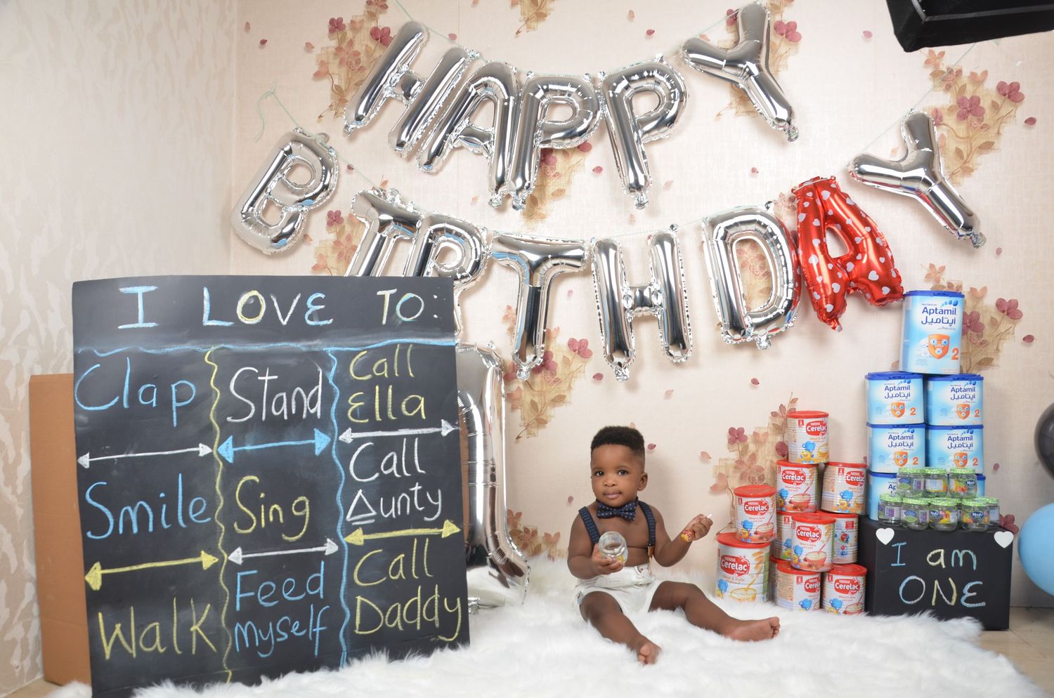 First birthday party setup with silver balloon letters and chalkboard milestone sign.