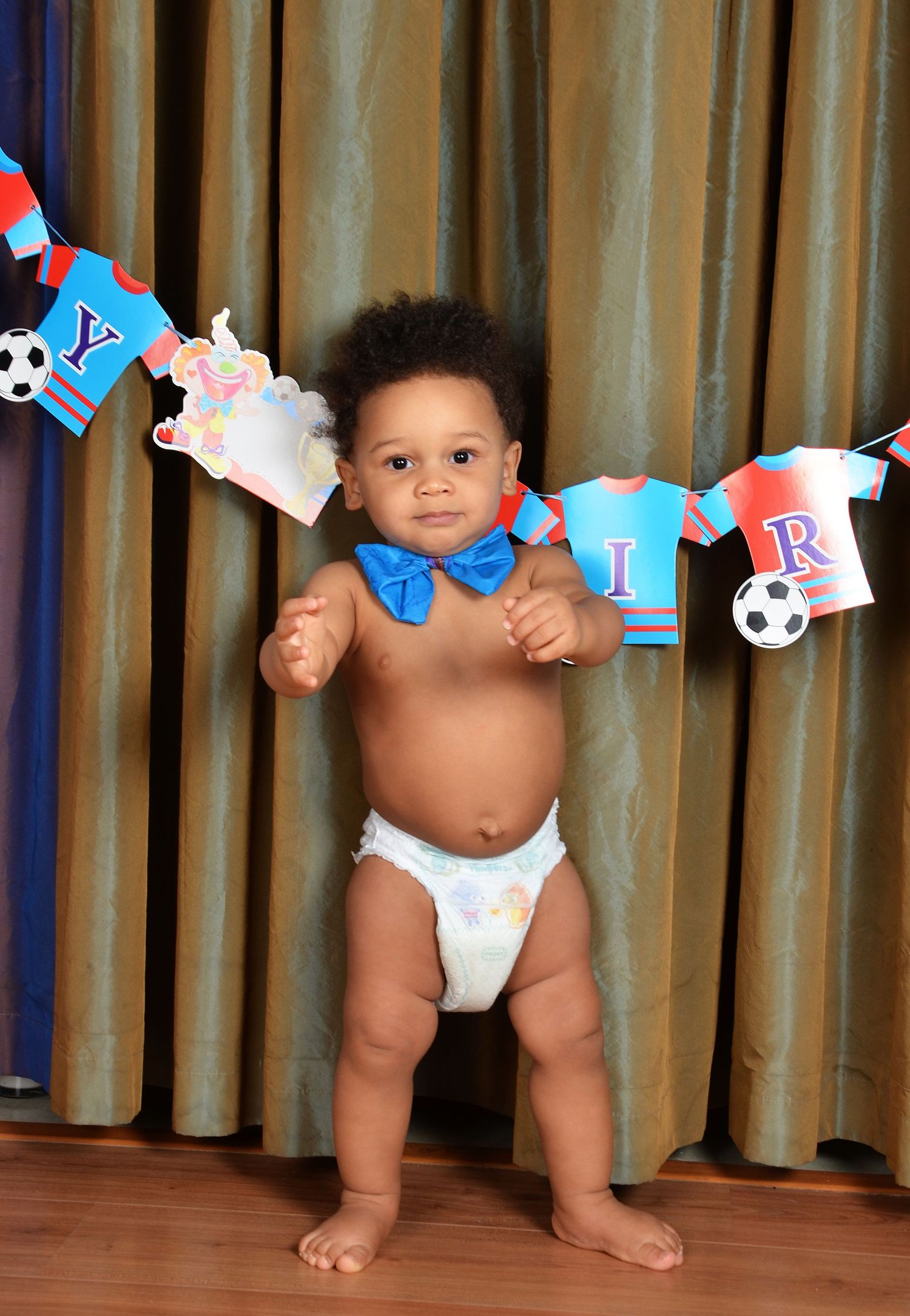 A baby wearing a blue bowtie stands in front of a soccer-themed birthday banner with tiny jerseys.