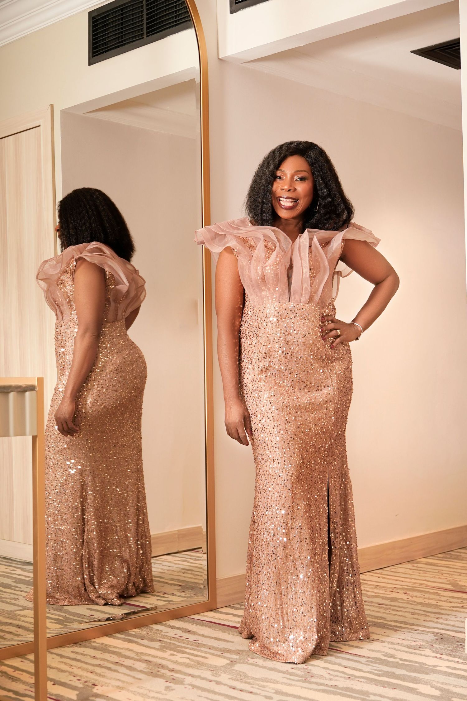 A glamorous model in a rose gold sequined gown poses elegantly by a full-length mirror.