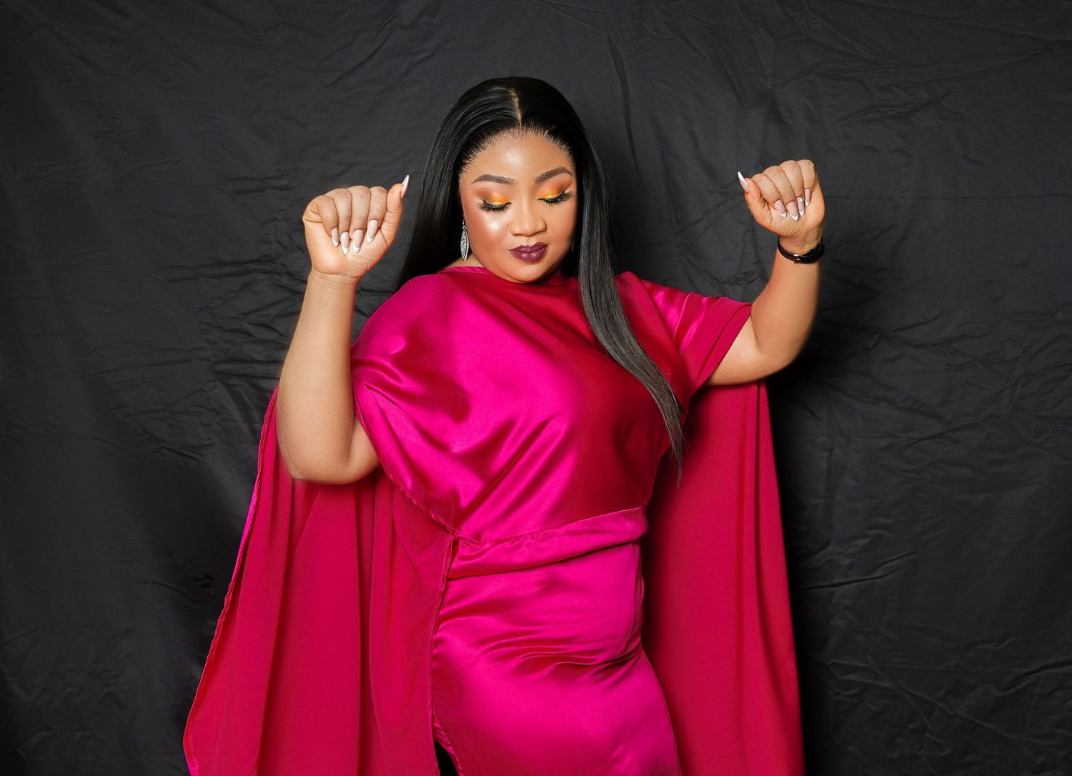A model in a flowing hot pink dress strikes a celebratory pose against black backdrop.