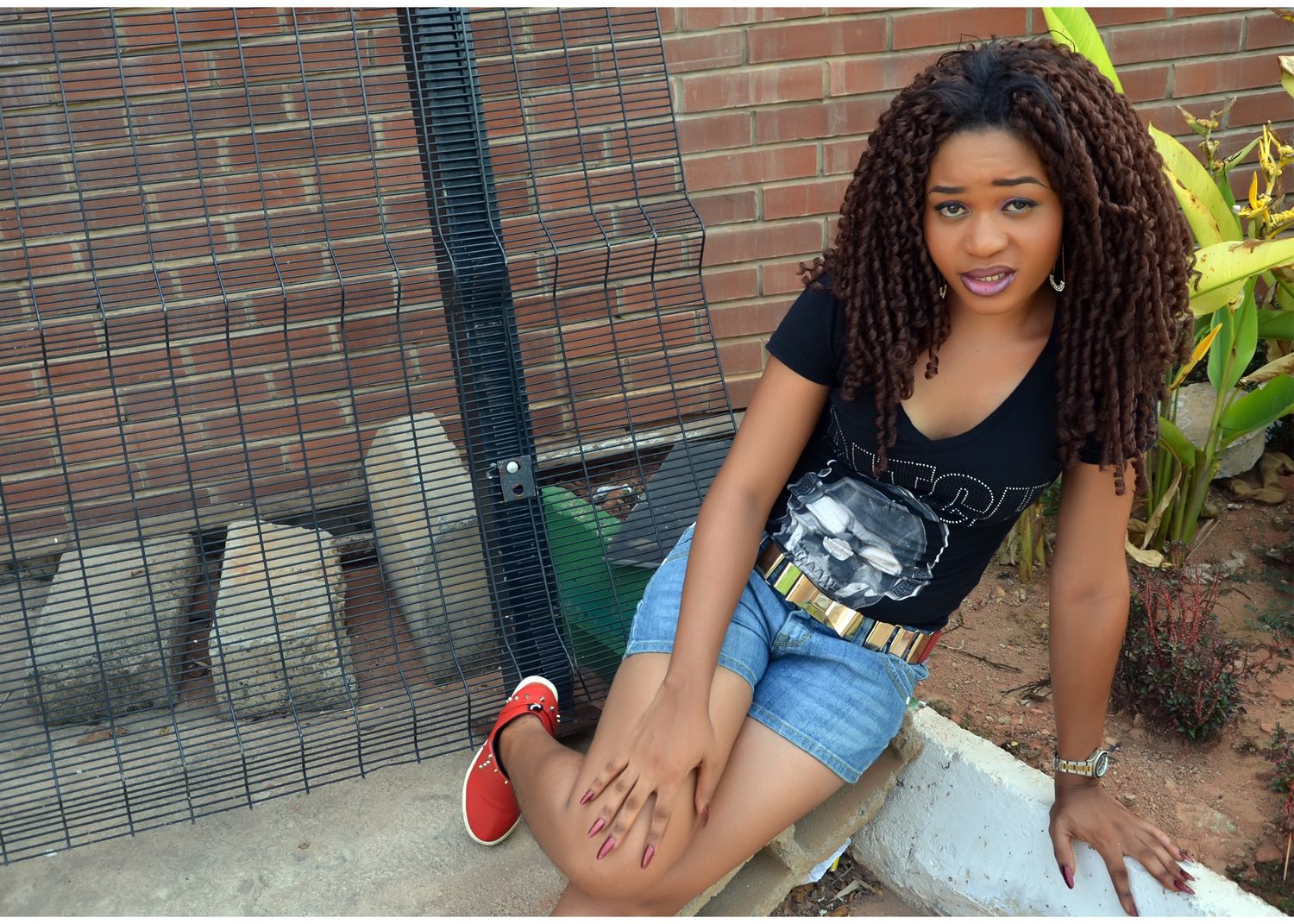 Woman in casual black top and denim shorts poses casually near brick wall and plants.