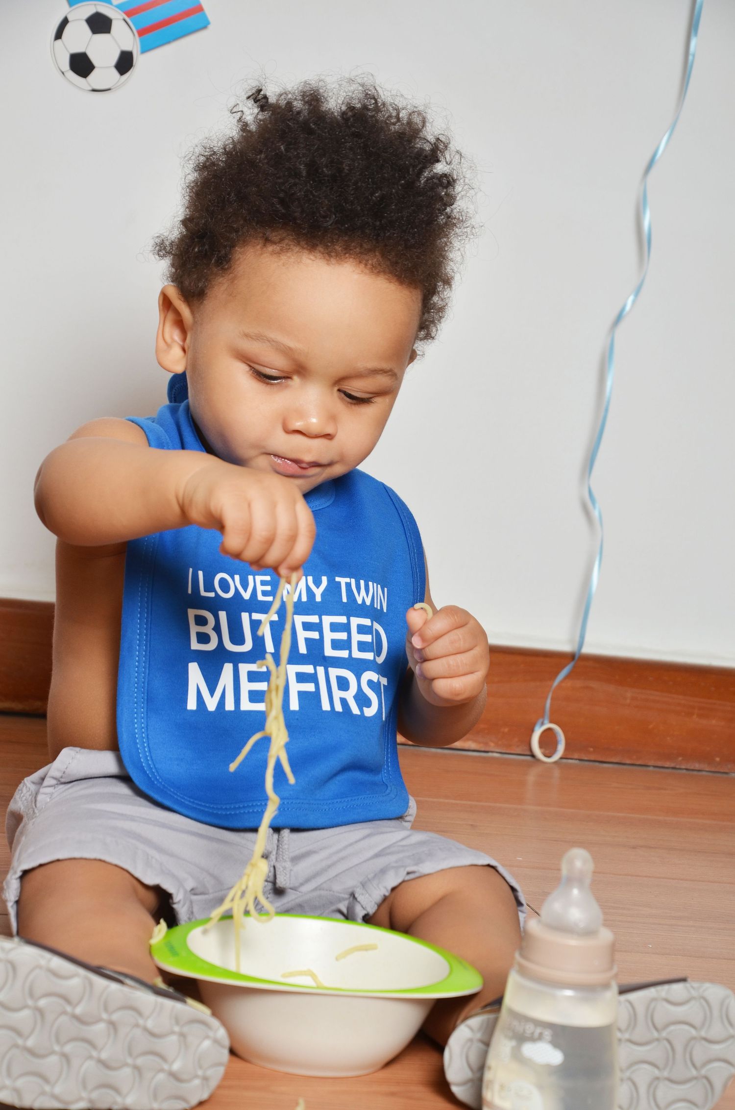 Baby wearing blue bib eating from plate with bottle nearby and streamers in background.