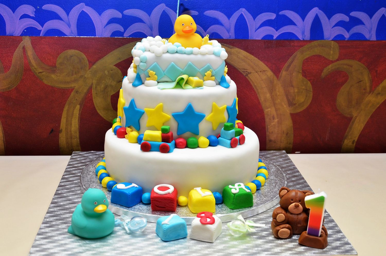 A colorful tiered birthday cake with rubber ducks, stars, and building blocks decorations.