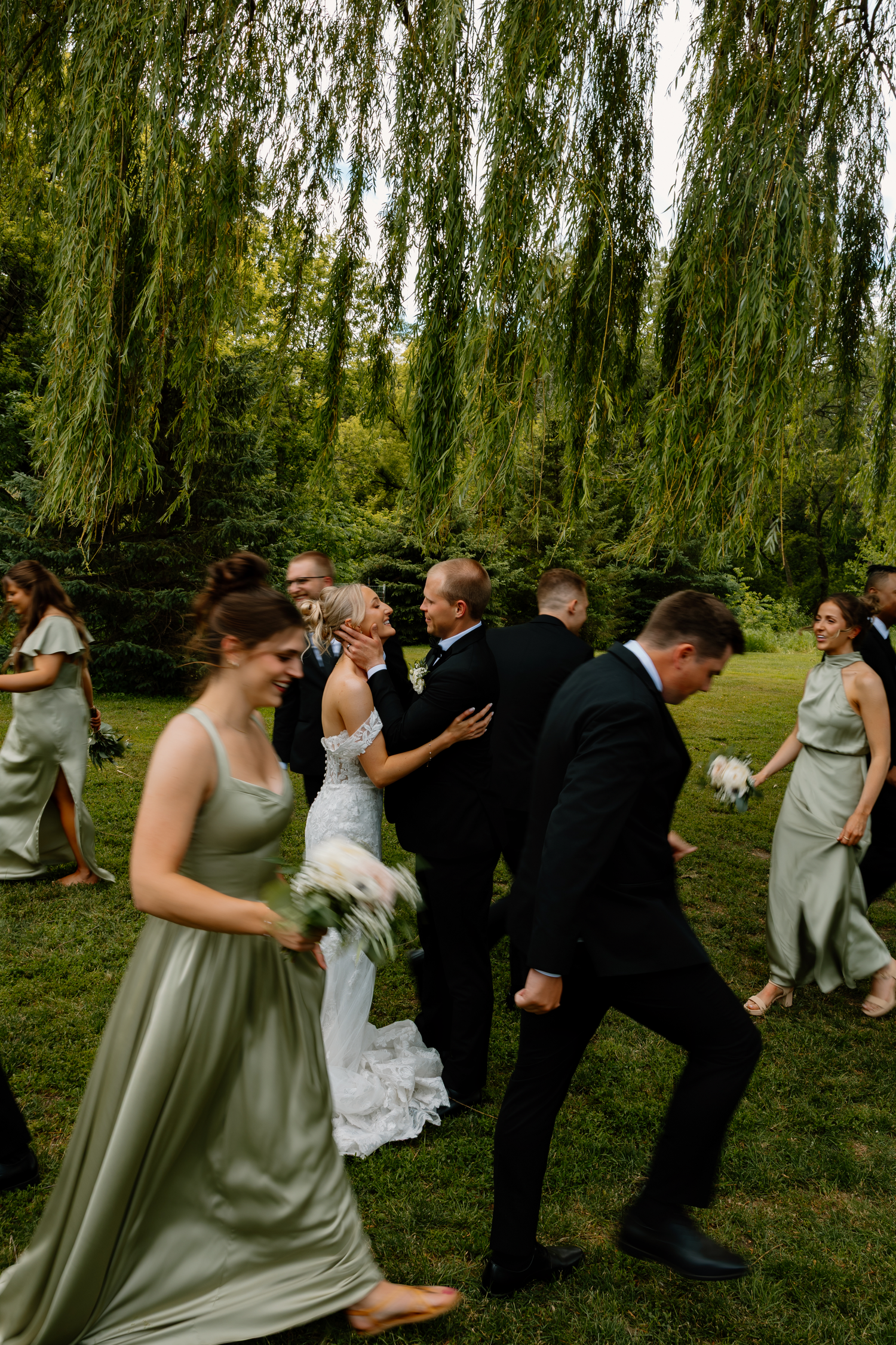 Wedding party laughs and celebrates outdoors beneath lush weeping willow trees after the ceremony.