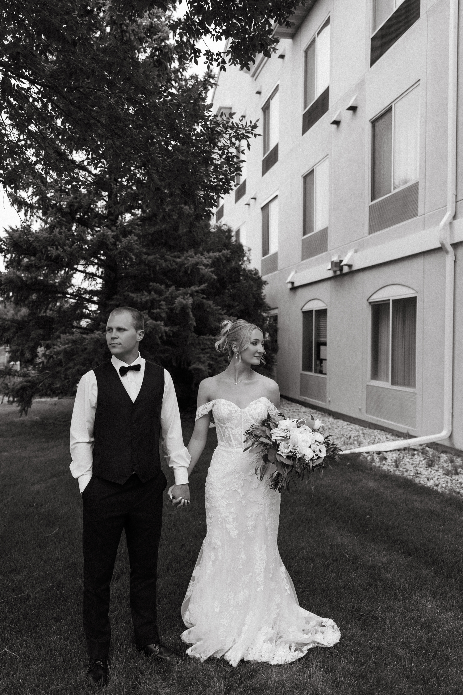 Black and white photo of a bride and groom holding hands outside a building, bride holding a floral bouquet.