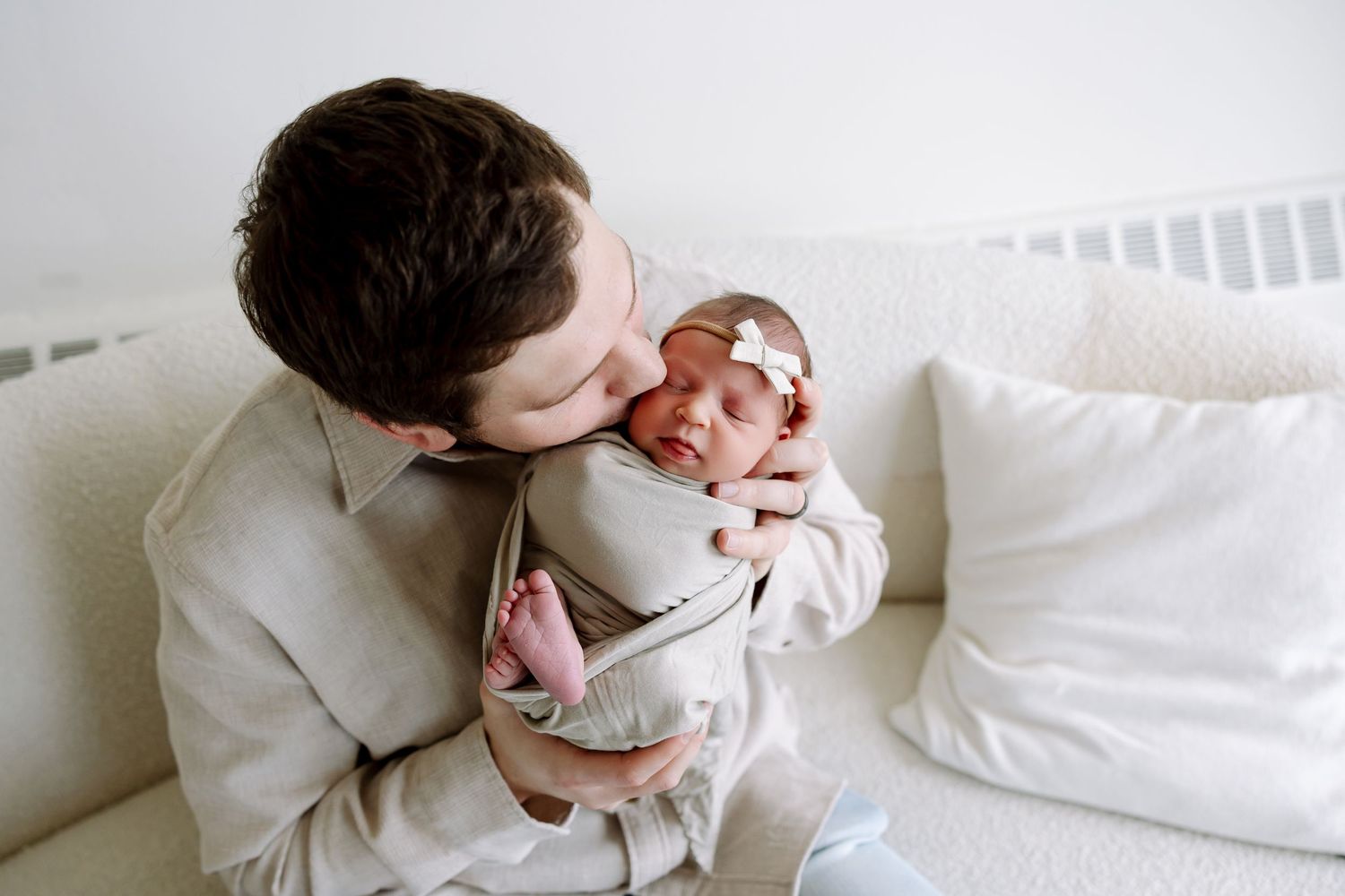 Parent and newborn baby sharing tender moments together on white bedding in soft natural light.