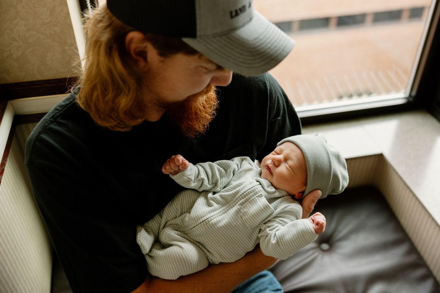 Baby in a white outfit rests in a parent's arms near a window.