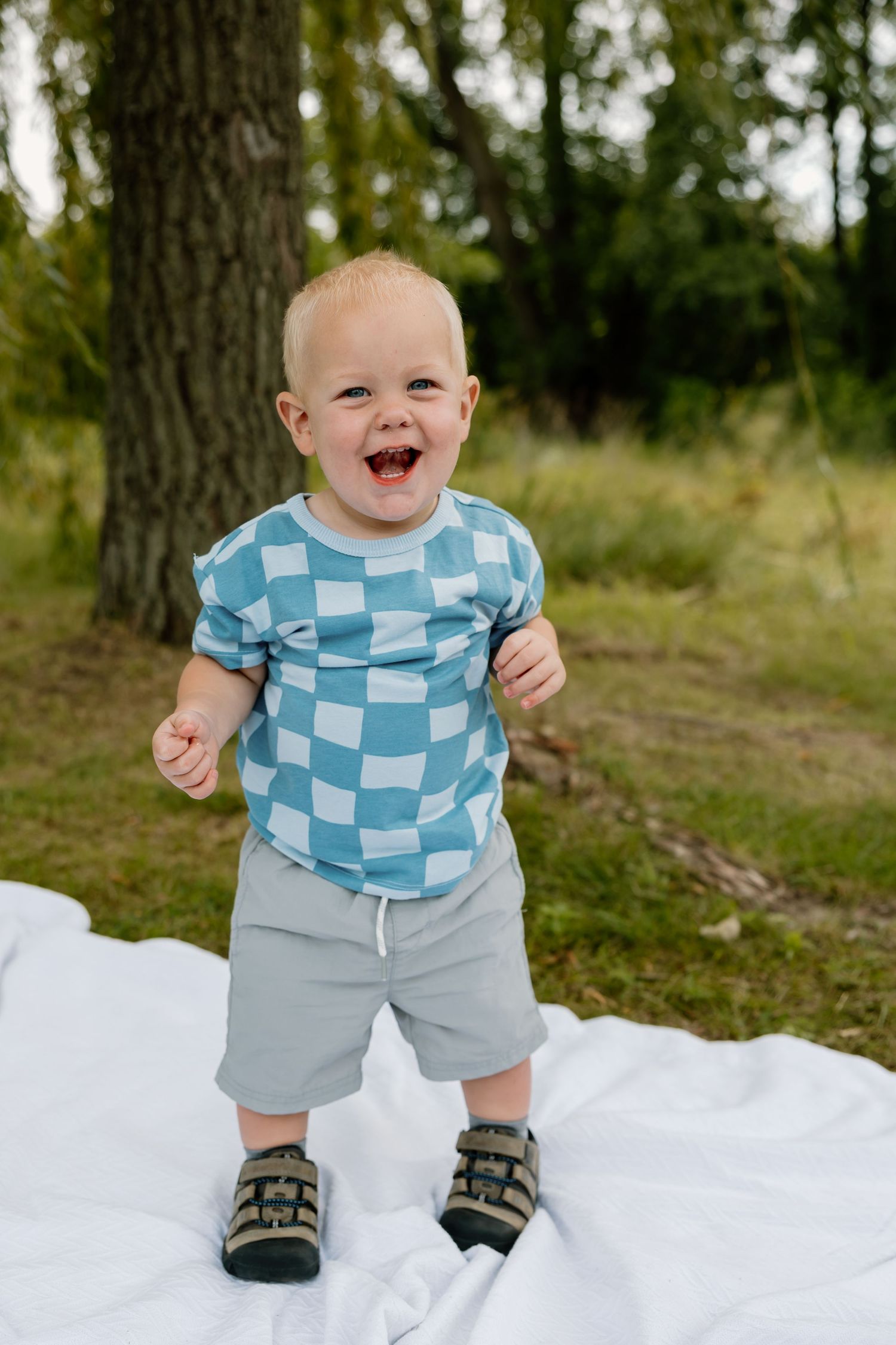 Joyful toddler in blue checkered shirt laughs while standing on white blanket under tree.