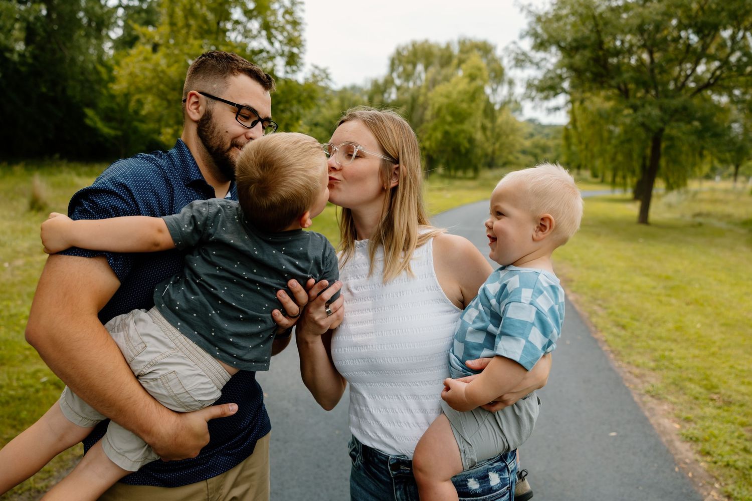 A family of four shares a playful moment together while standing on a paved path surrounded by green trees.