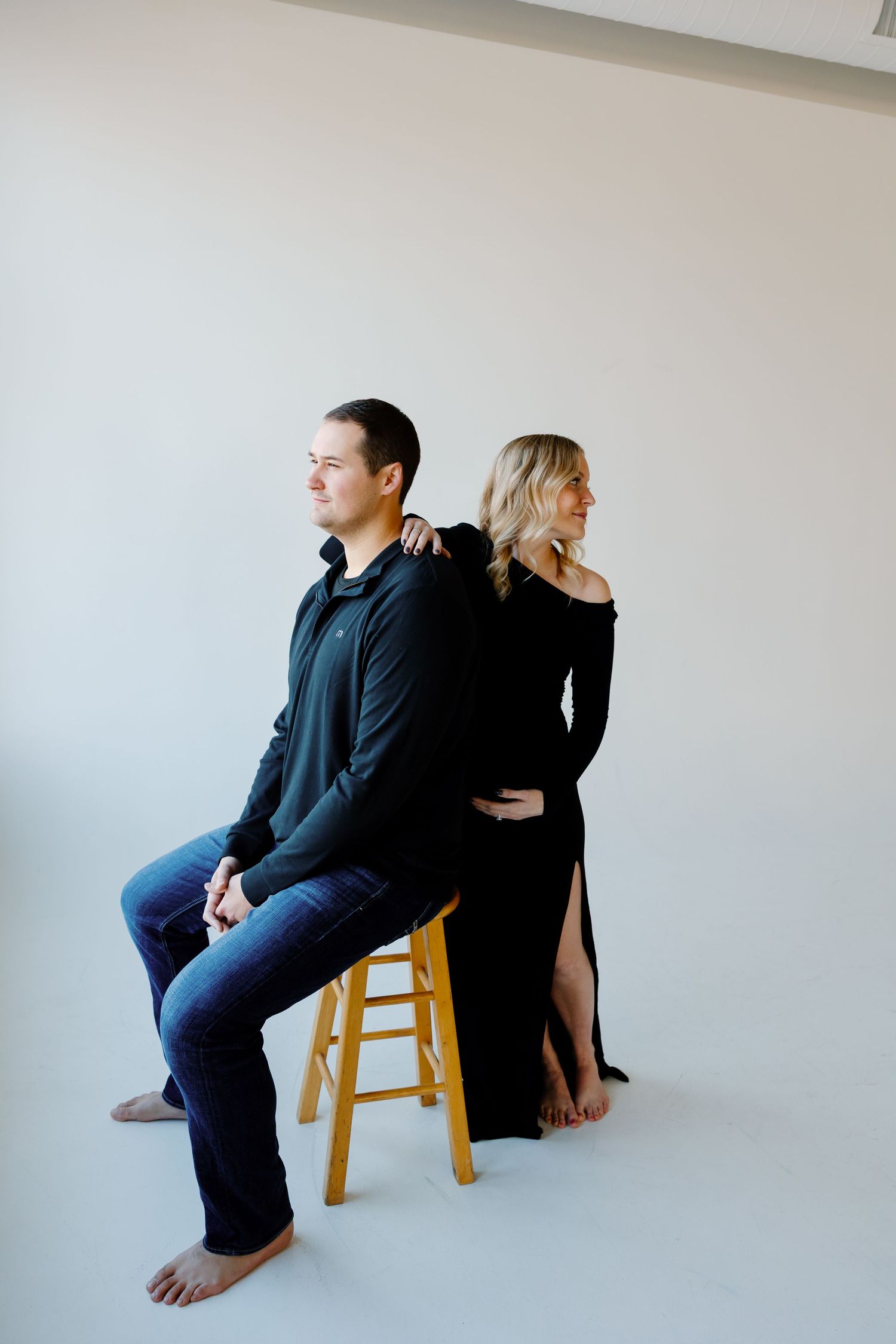 Two people sitting back-to-back on a wooden stool against a minimalist white wall. Riley Ro Photo Sioux Falls, SD