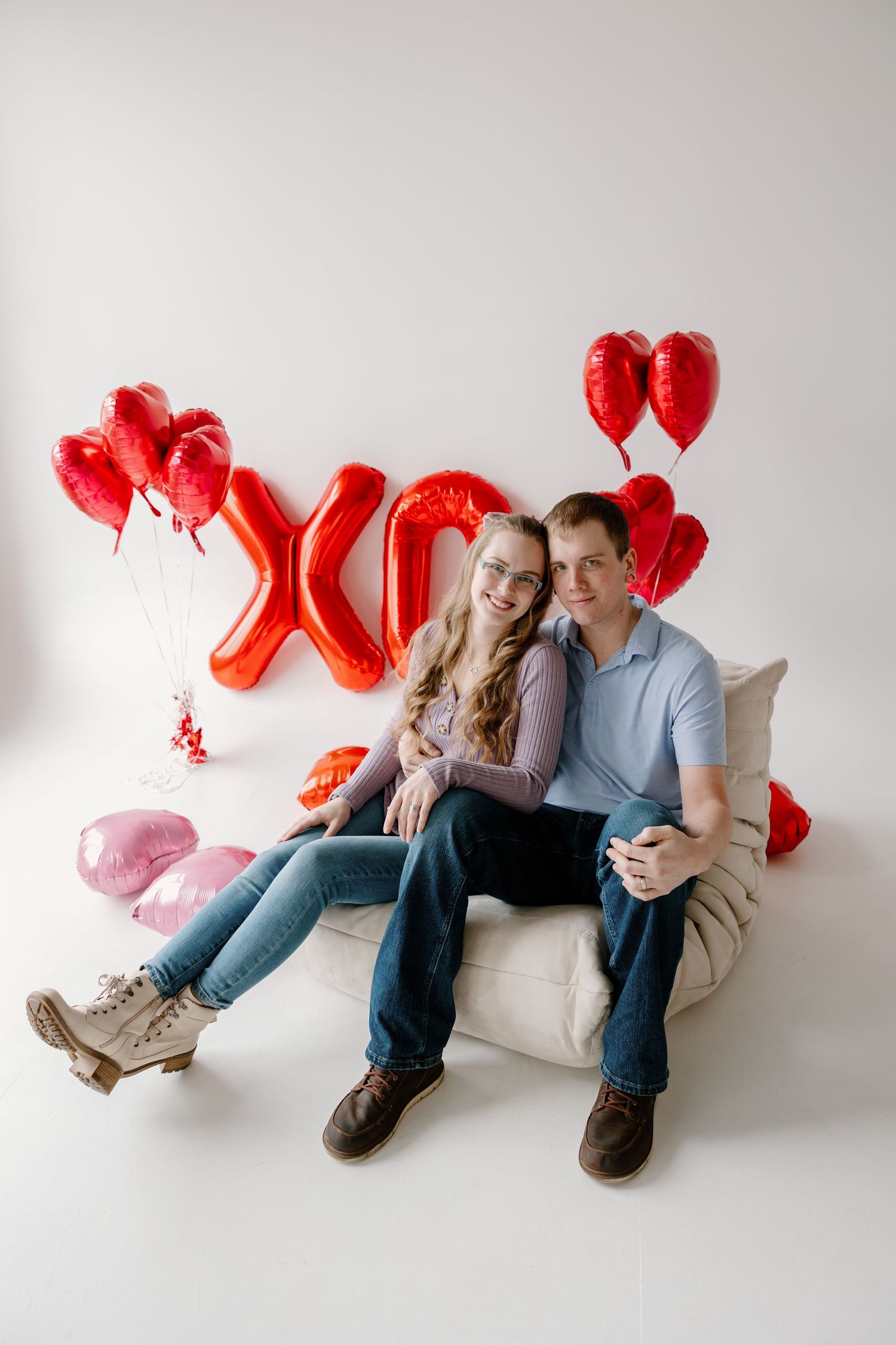 Couple sits on white couch with red heart balloons and XO decorations for Valentine's Day photo shoot.