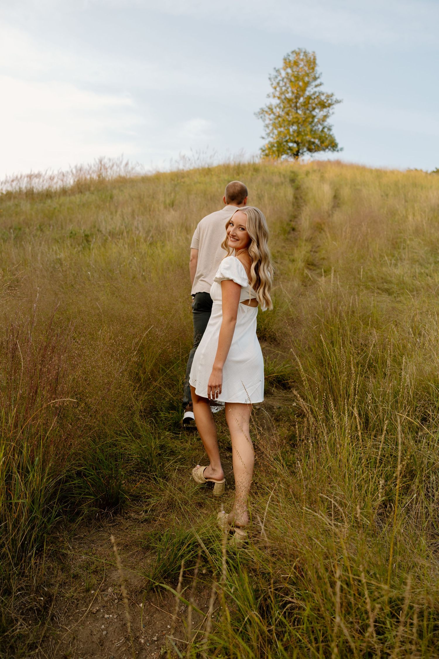 A couple walking up a grassy hillside together at sunset with a lone tree on the horizon.