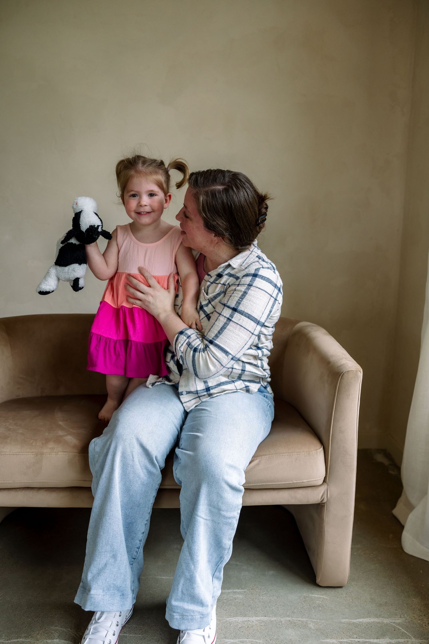 A woman kisses a smiling toddler girl in a pink dress holding a stuffed animal while sitting on a beige sofa.