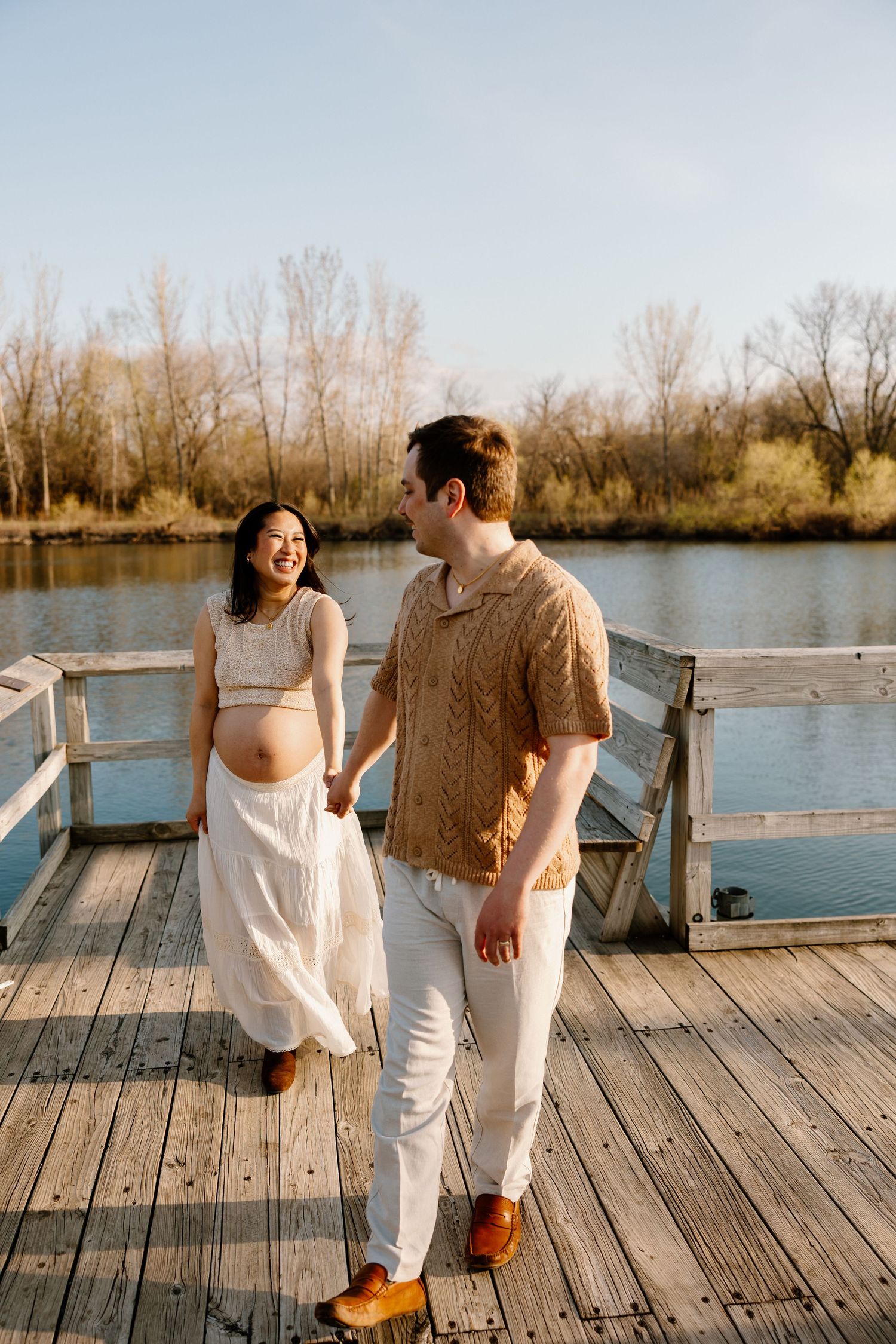 A couple shares a lighthearted moment on a wooden dock with autumn trees in the background.