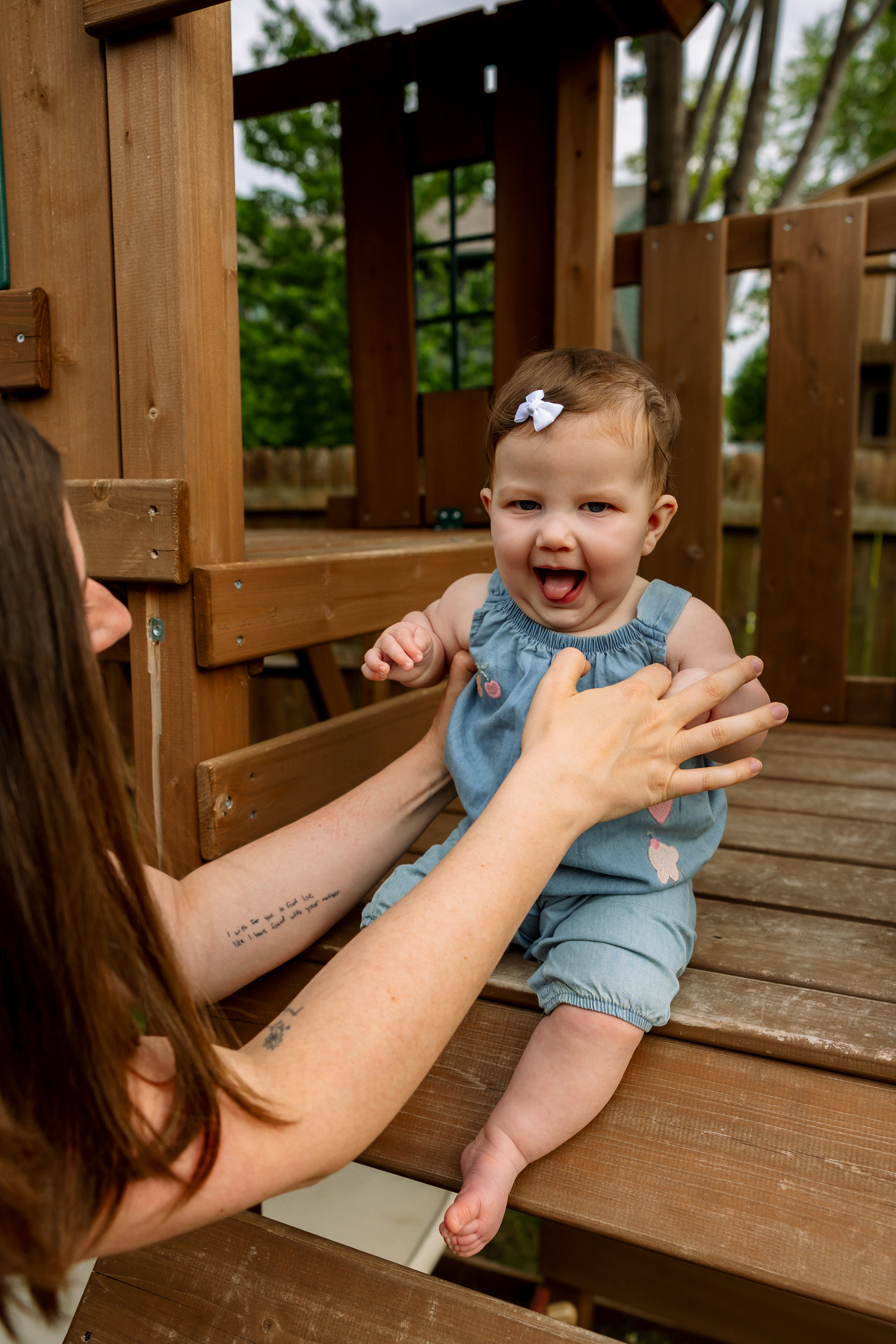 A laughing baby in a blue romper sits on a wooden playset while a mother reaches out to her outdoors.