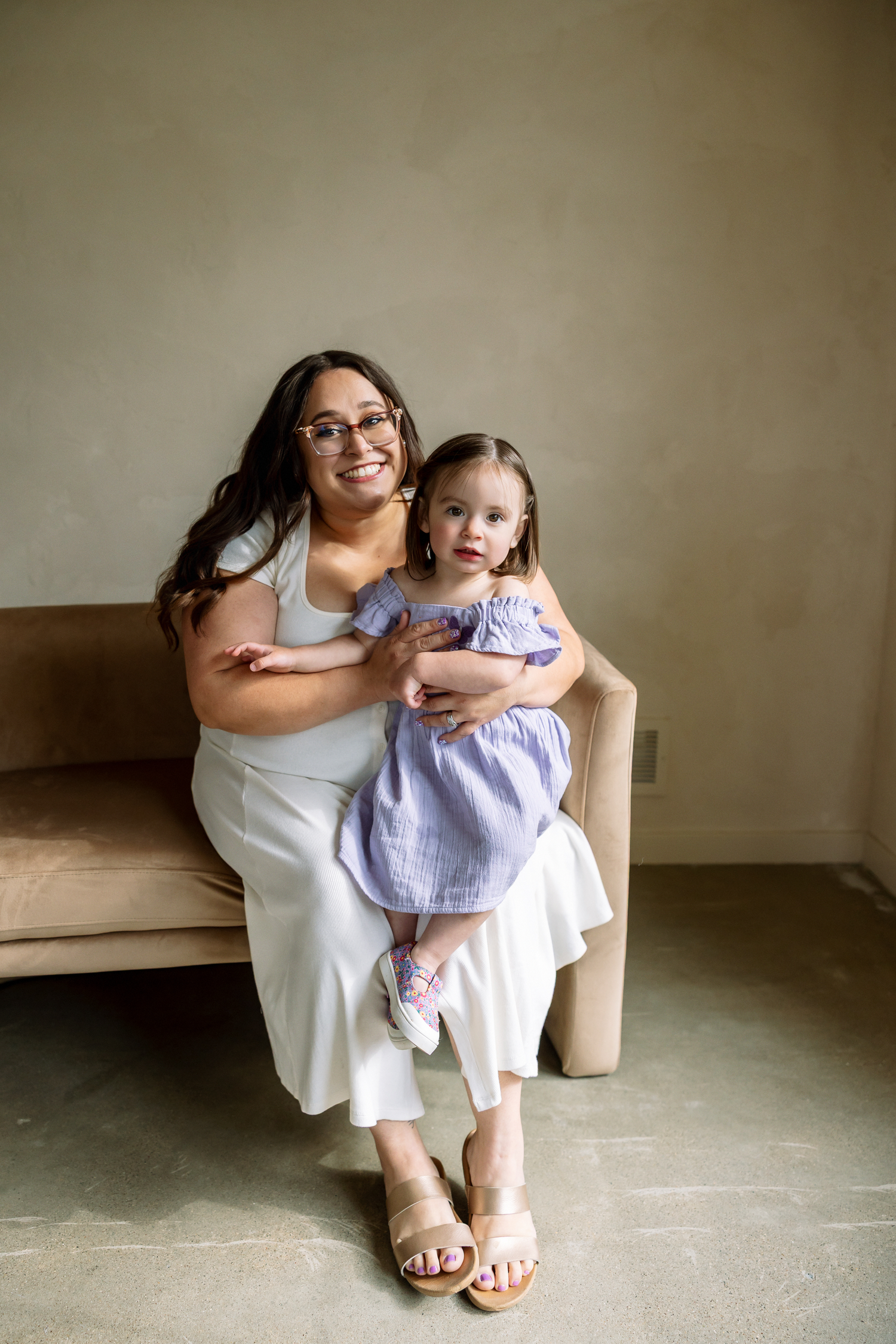 A smiling woman in white sits on a beige chair holding a young girl in a blue striped dress.