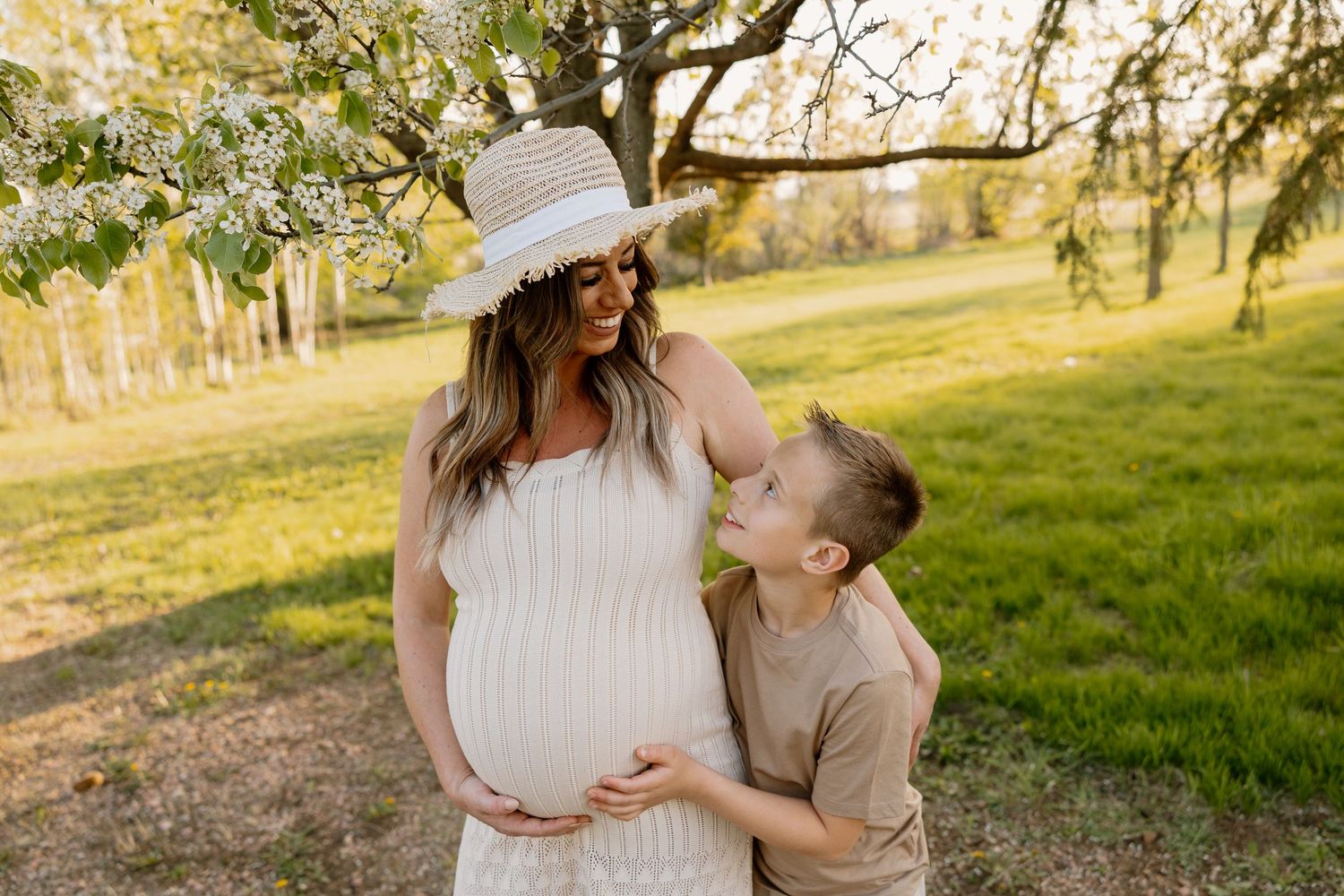 An expecting mother in a white dress and sun hat shares a joyful moment with her child in a sunlit field.