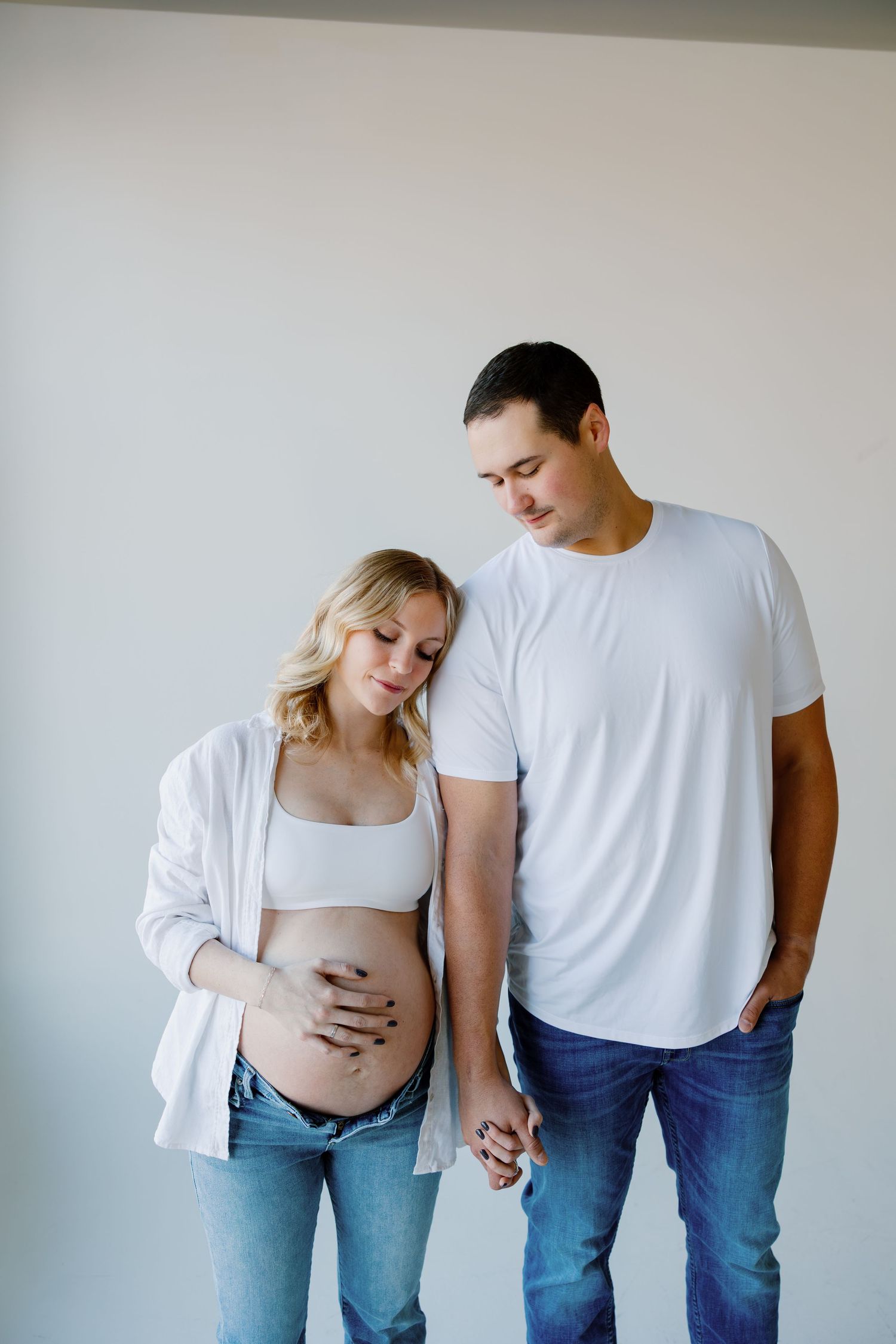 A couple in white shirts and jeans pose together while expecting a baby in a minimalist studio setting. Riley Ro Photo Sioux Falls, SD