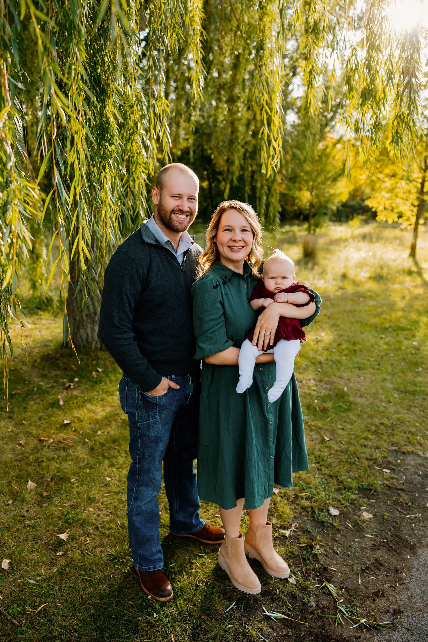 A smiling family of three poses outdoors under a willow tree, with parents holding their baby in a sunny green park.