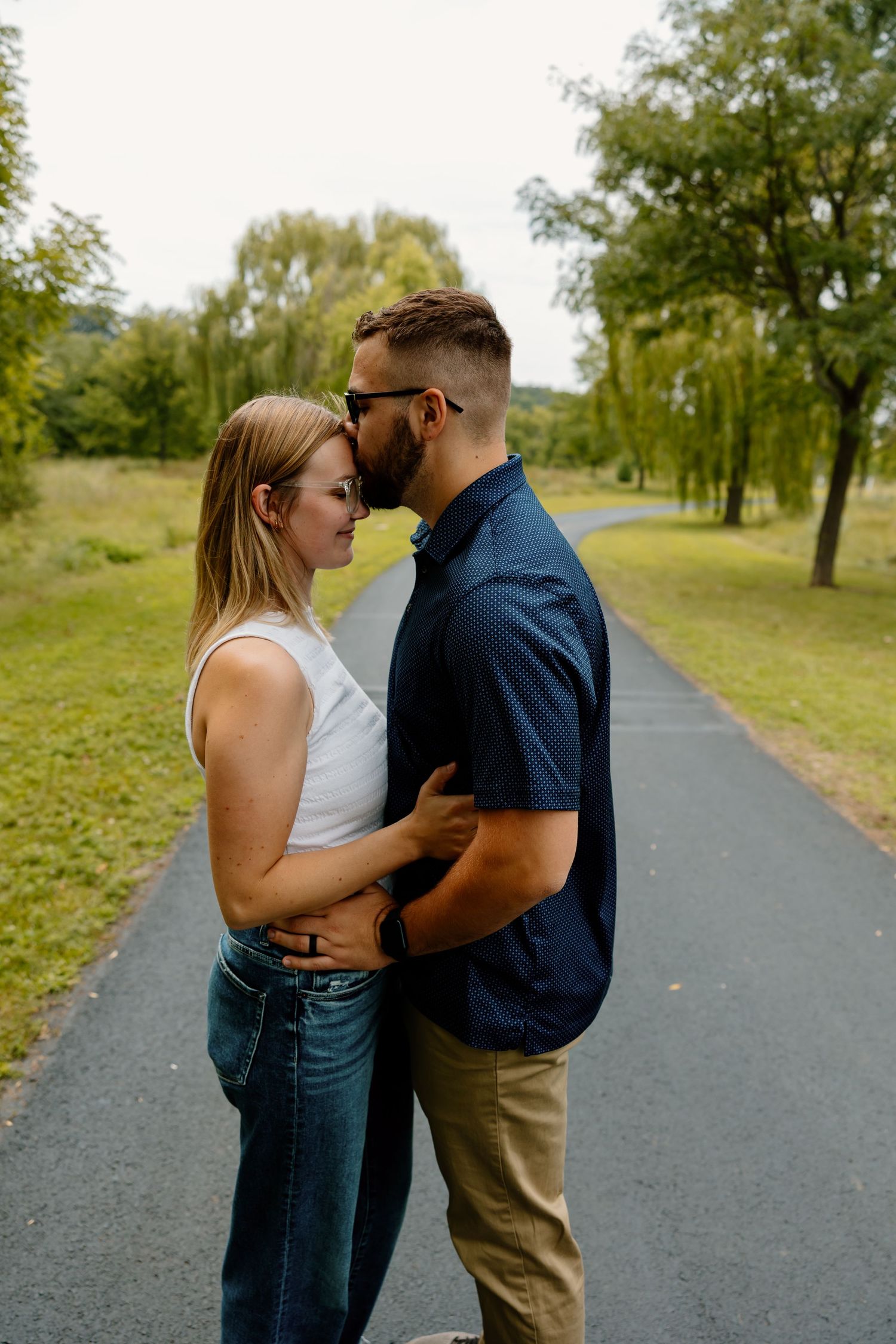 A couple shares romantic moments together while standing on a paved path surrounded by green grass and trees.