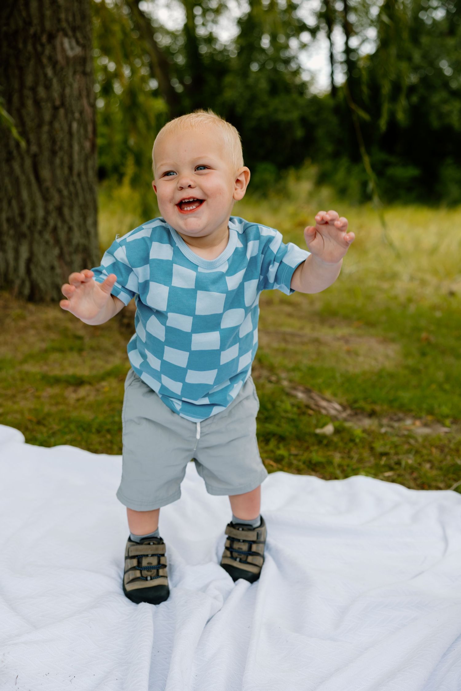 Laughing toddler in blue checkered shirt and shorts standing on white blanket outdoors.