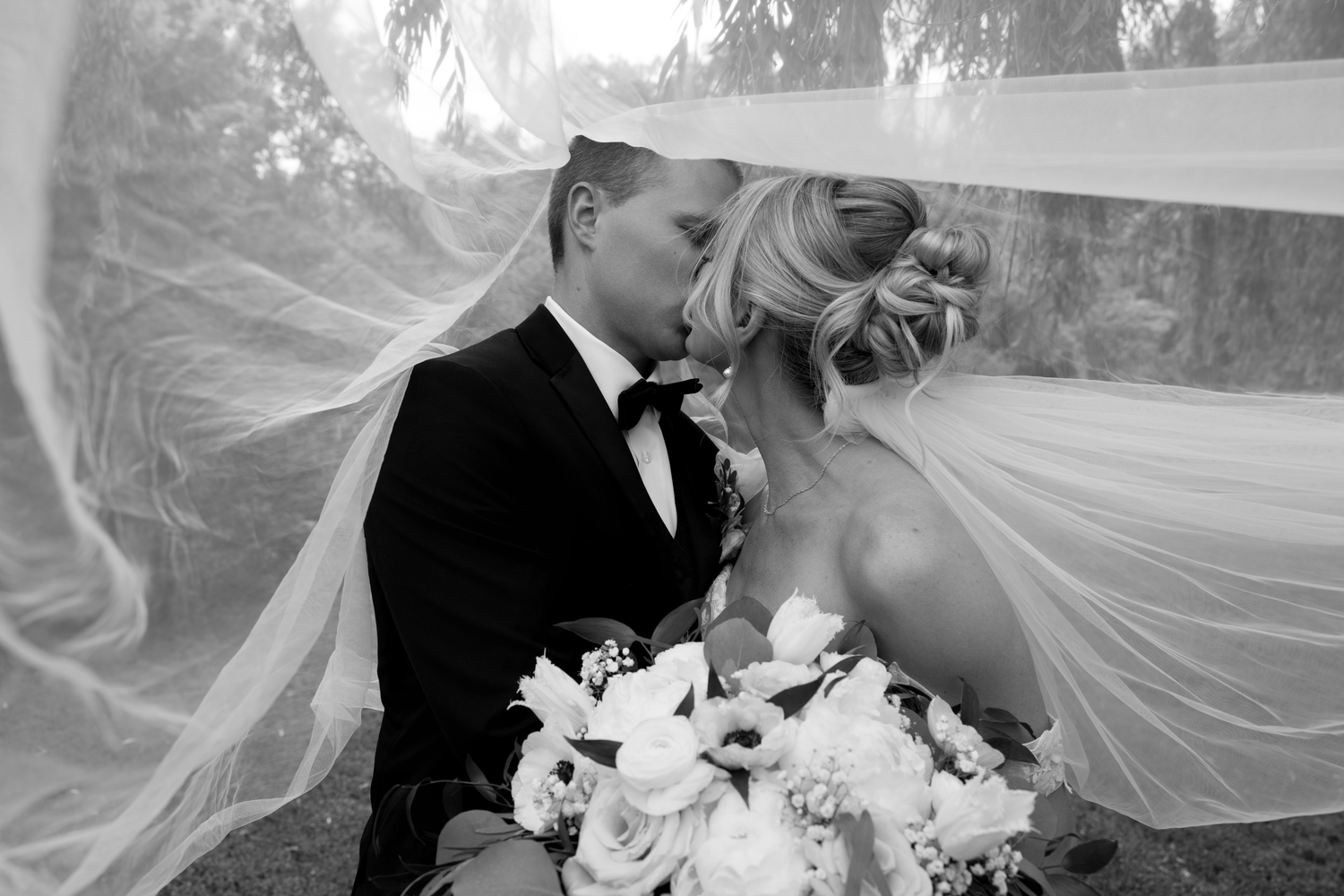 Black and white wedding photo of bride and groom kissing under flowing veil, bride holding white floral bouquet.