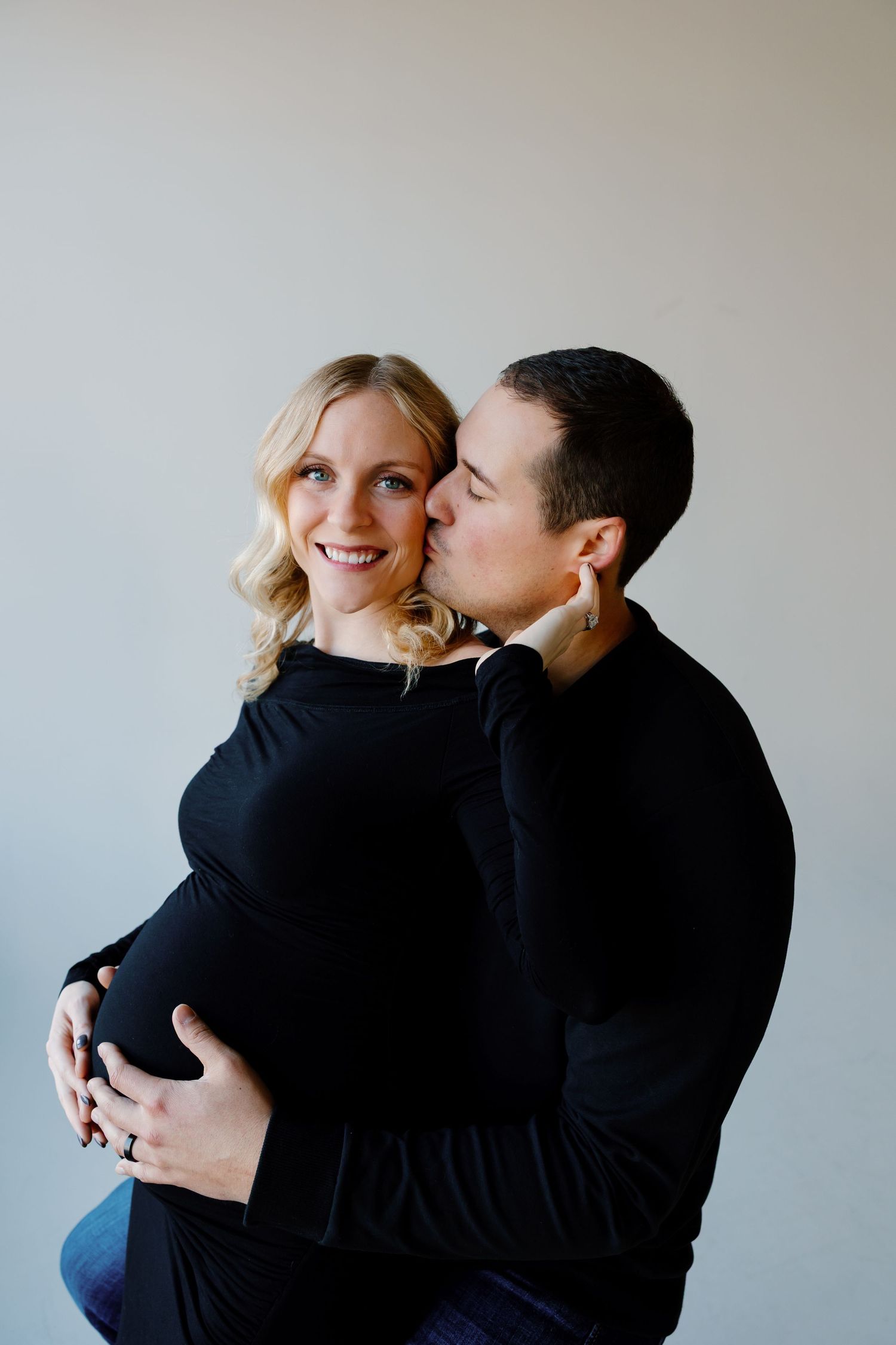A couple wearing black outfits embraces intimately against a light gray wall in a series of romantic candid photos.