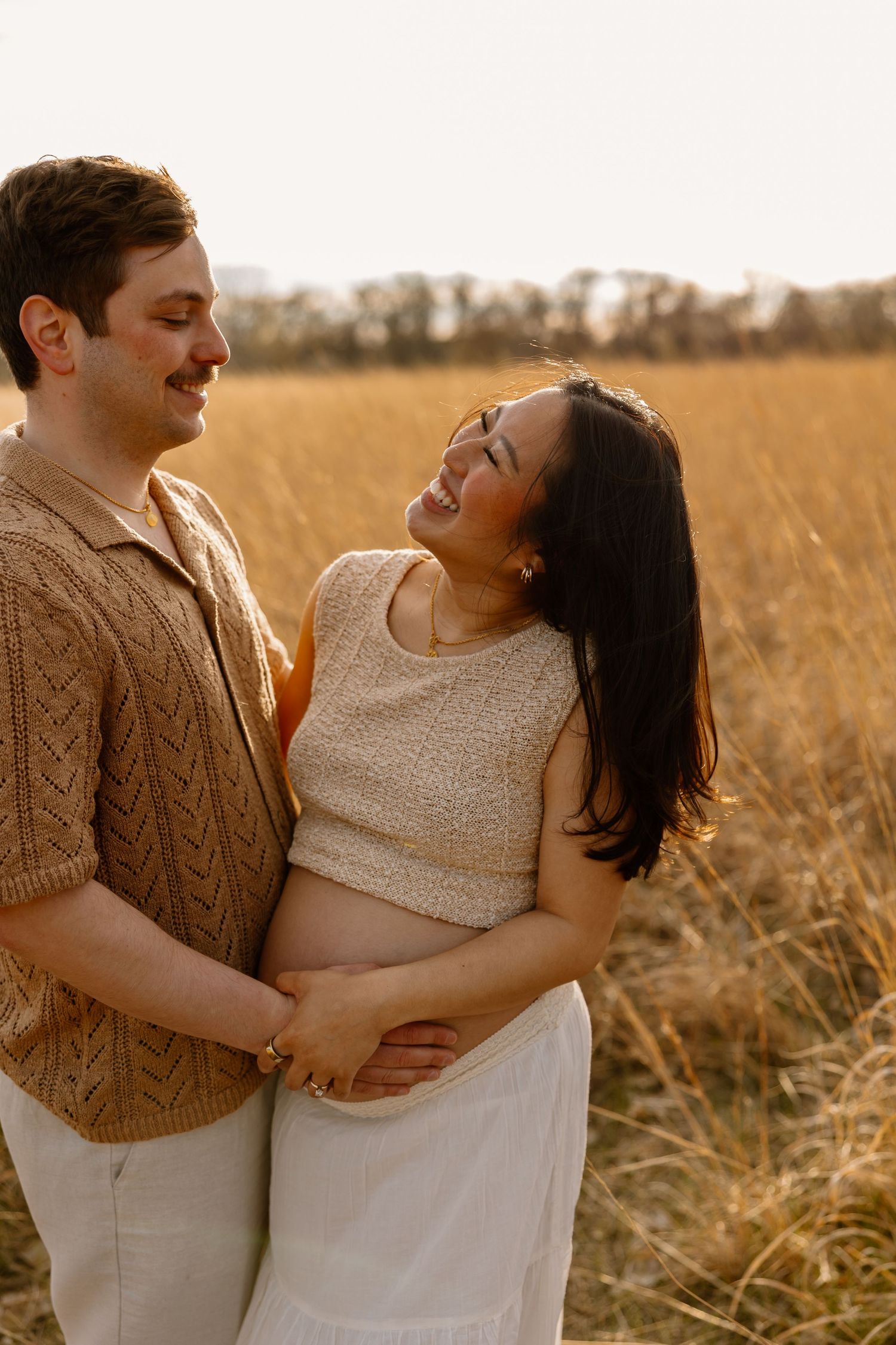 A couple shares an intimate moment during their maternity photoshoot in a sunlit field of tall grass.