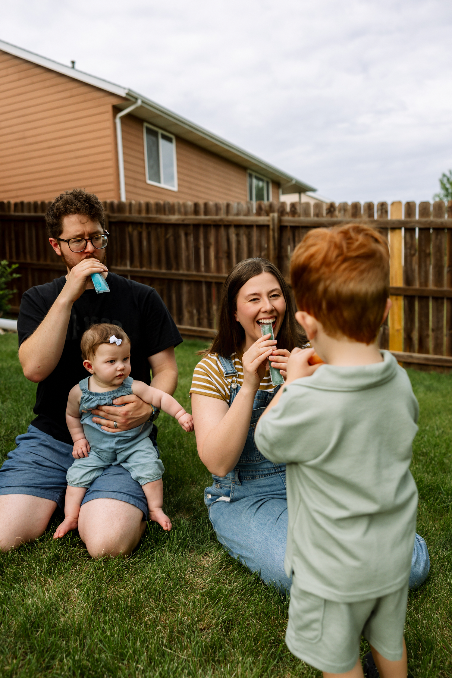 Family of four sitting on grass in backyard enjoying colorful popsicles together on a cloudy day.