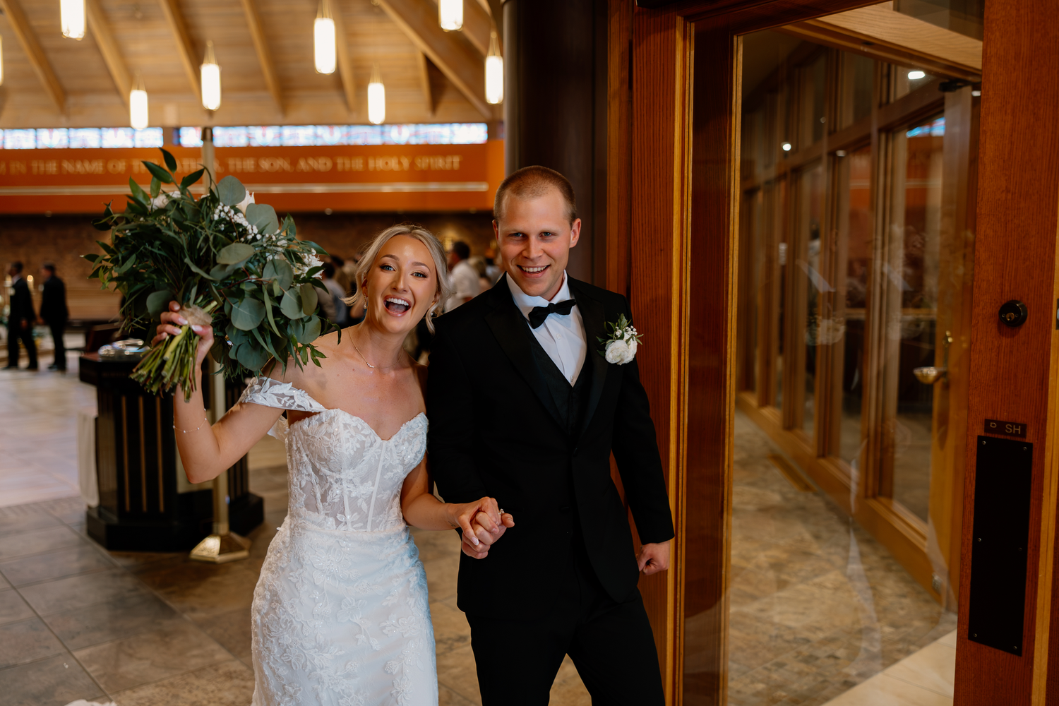 Joyful newlyweds exit church holding hands, bride in lace gown with bouquet, groom in black tuxedo, celebrating wedding day.