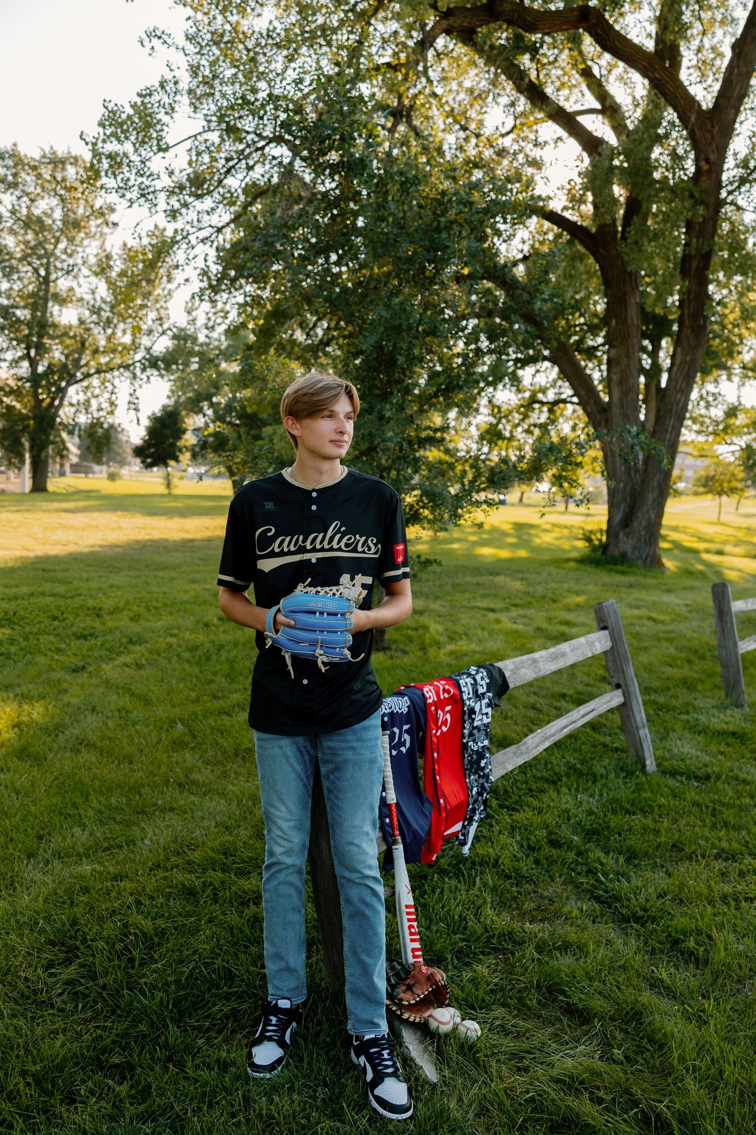 A person in jeans and a graphic t-shirt stands near a wooden fence in a sunny field.