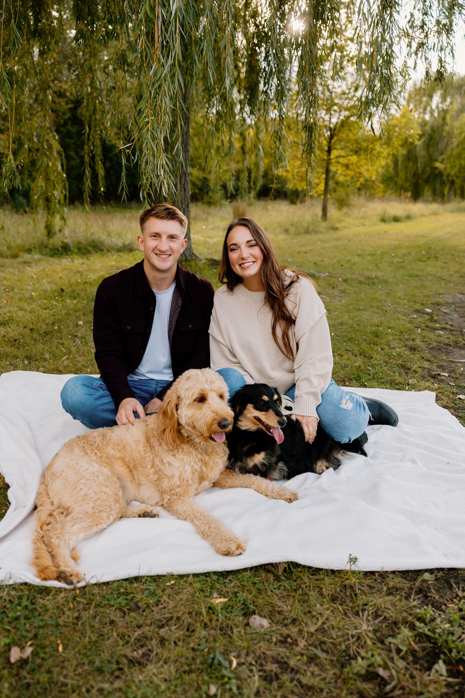 A couple sits on a white blanket under a willow tree with their two dogs during an autumn outdoor photoshoot. Riley Ro Photo Sioux Falls, SD