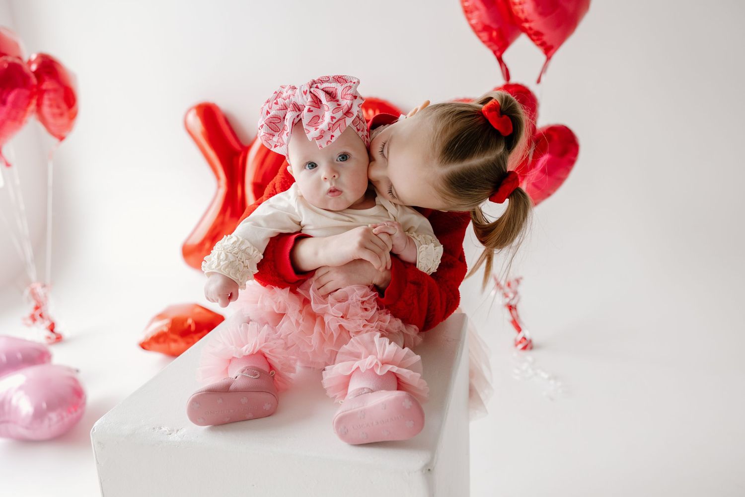 A person sits with a baby surrounded by pink and red Valentine's Day decorations on a white surface.