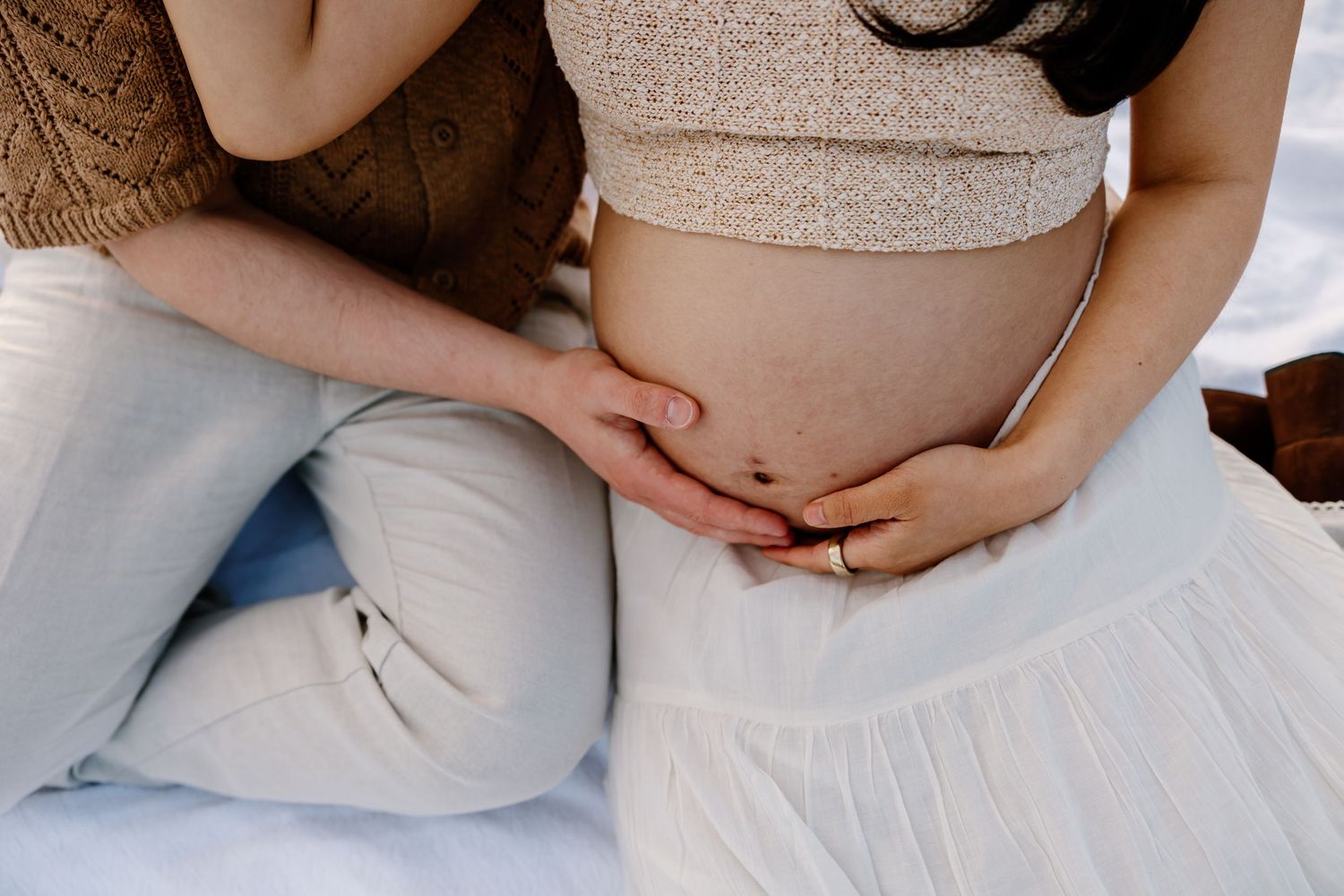 Close up of pregnant belly being cradled in loving hands while sitting on white fabric.