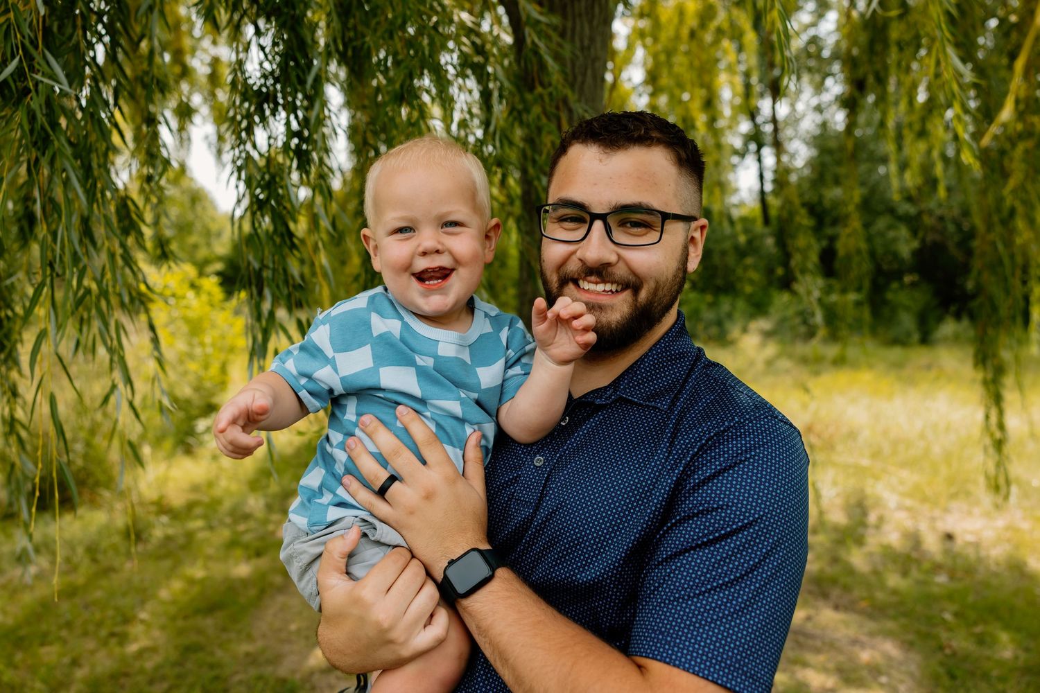 Joyful parent in navy blue shirt cuddling and playing with baby under weeping willow trees in park setting.