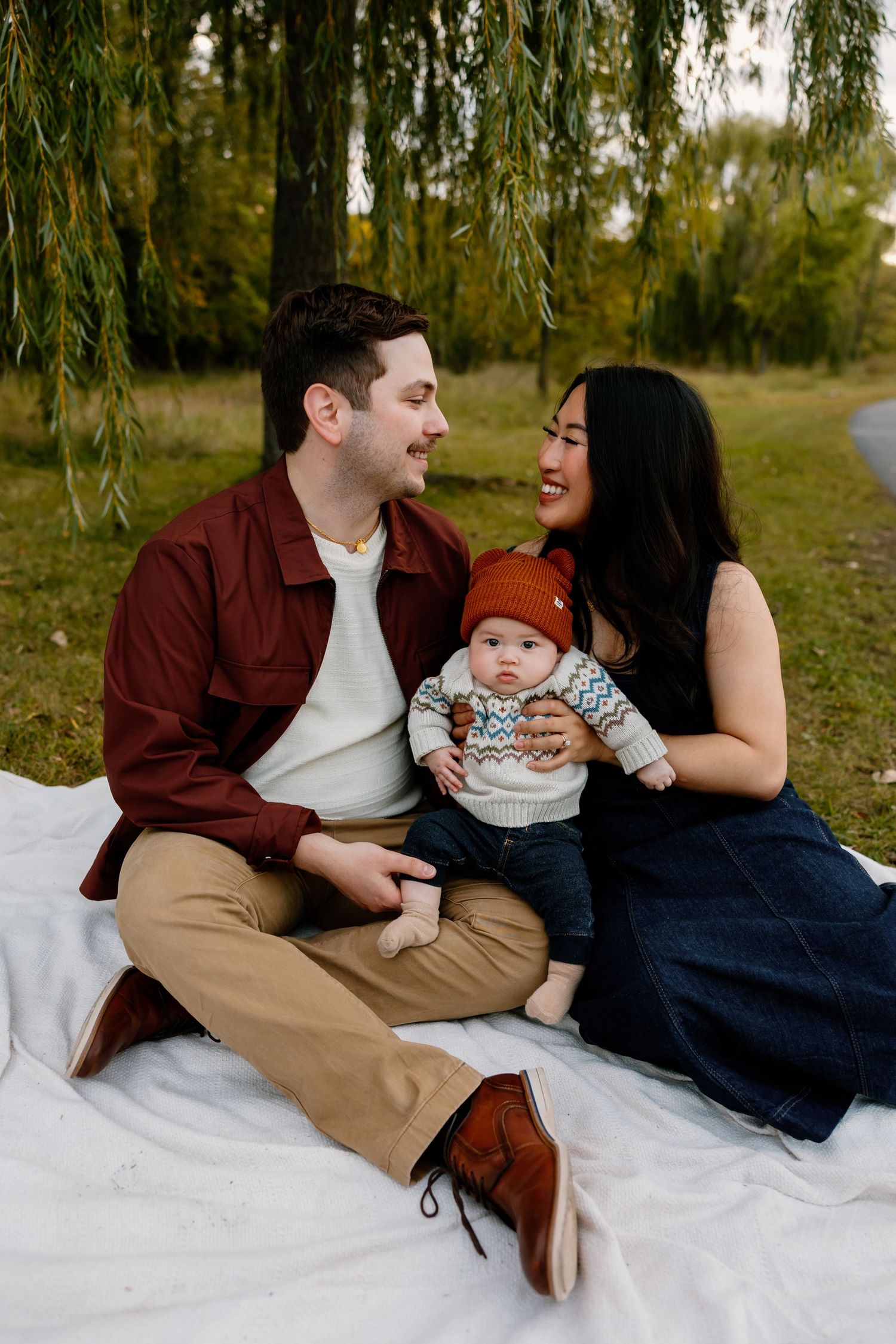 A young family sits on a white blanket in a park, parents smiling at each other with their baby in a rust knit hat.