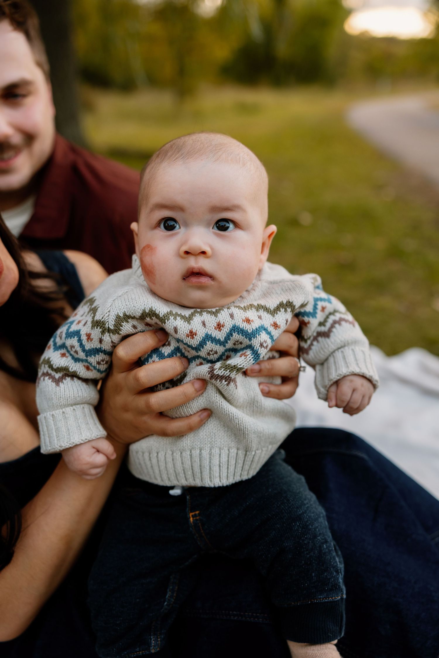 Adorable baby in a fair isle sweater being held by parents outdoors in a park during golden hour.