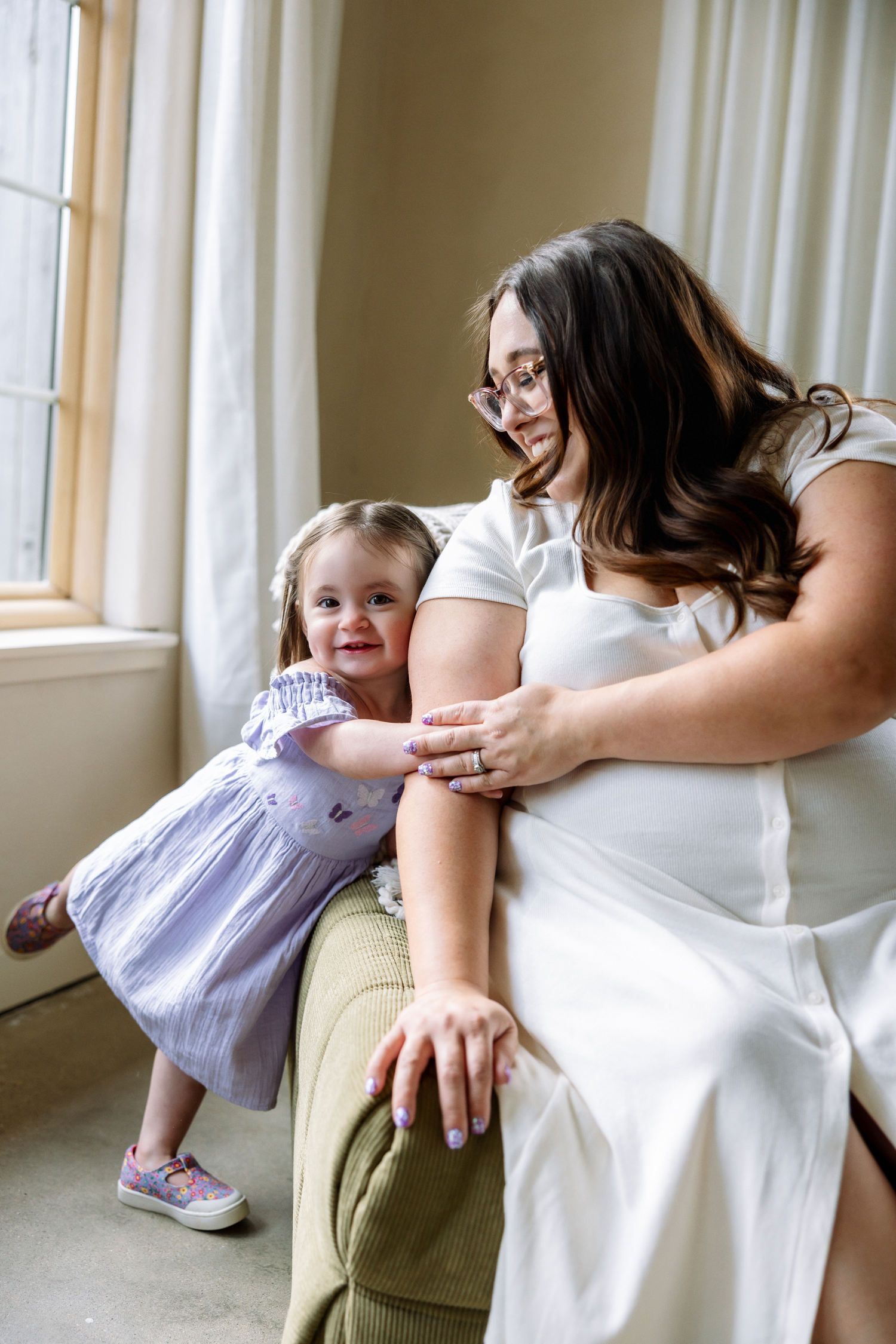 Pregnant woman in white dress smiling at toddler girl in lavender dress leaning on chair by window.