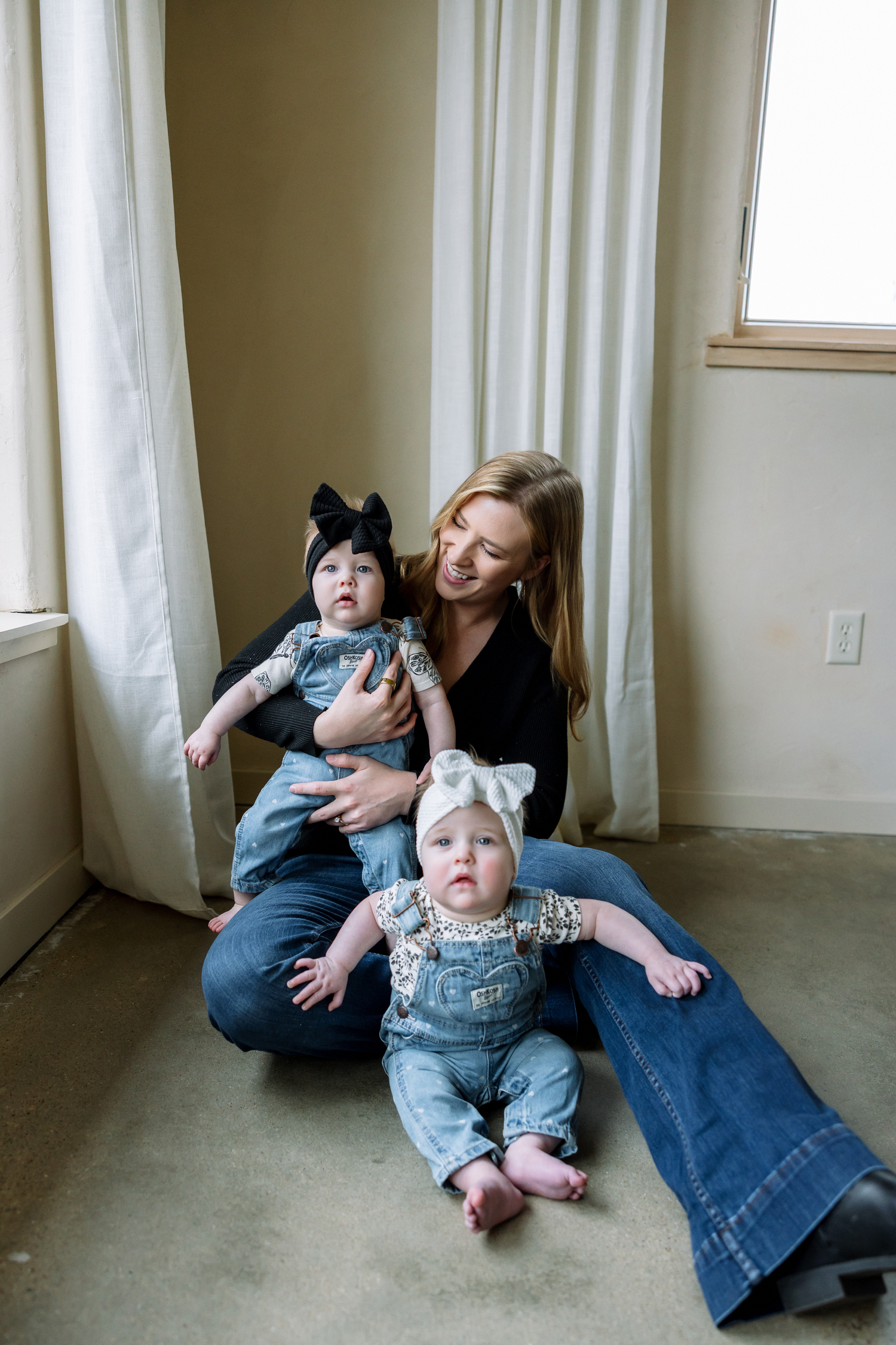 A smiling mother in a black top and jeans sits on the floor holding two baby girls in denim overalls and bow headbands.
