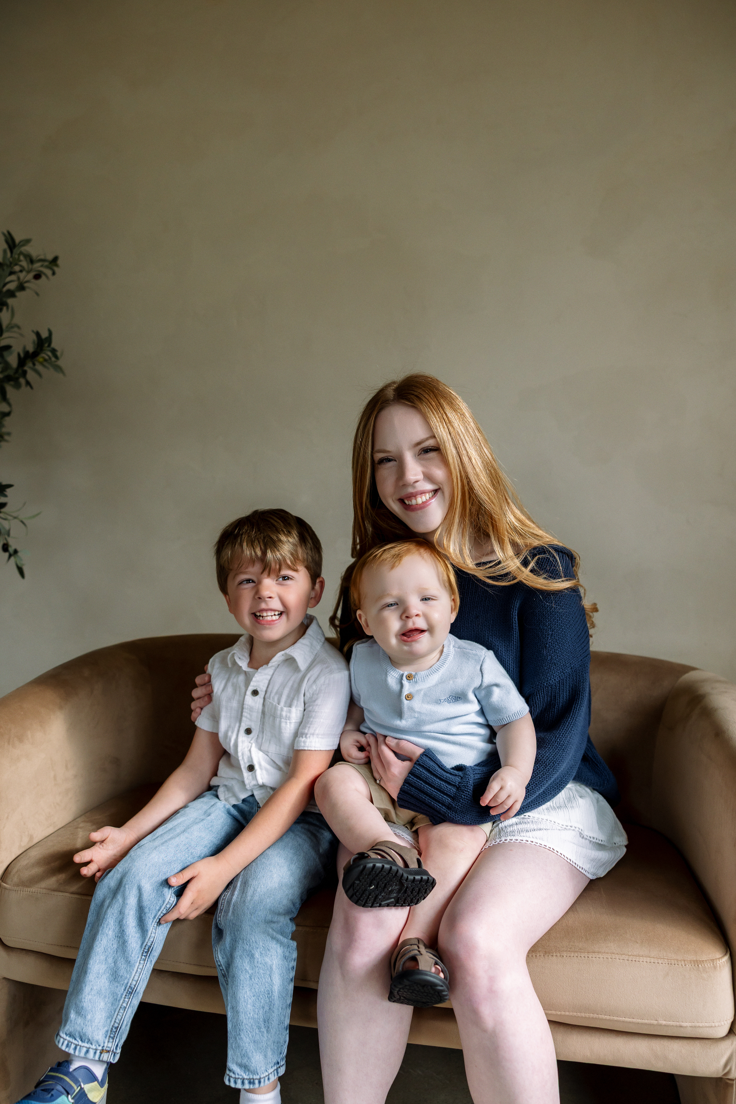 A smiling teenage girl with red hair sits on a beige sofa holding a baby while a young boy sits beside her.
