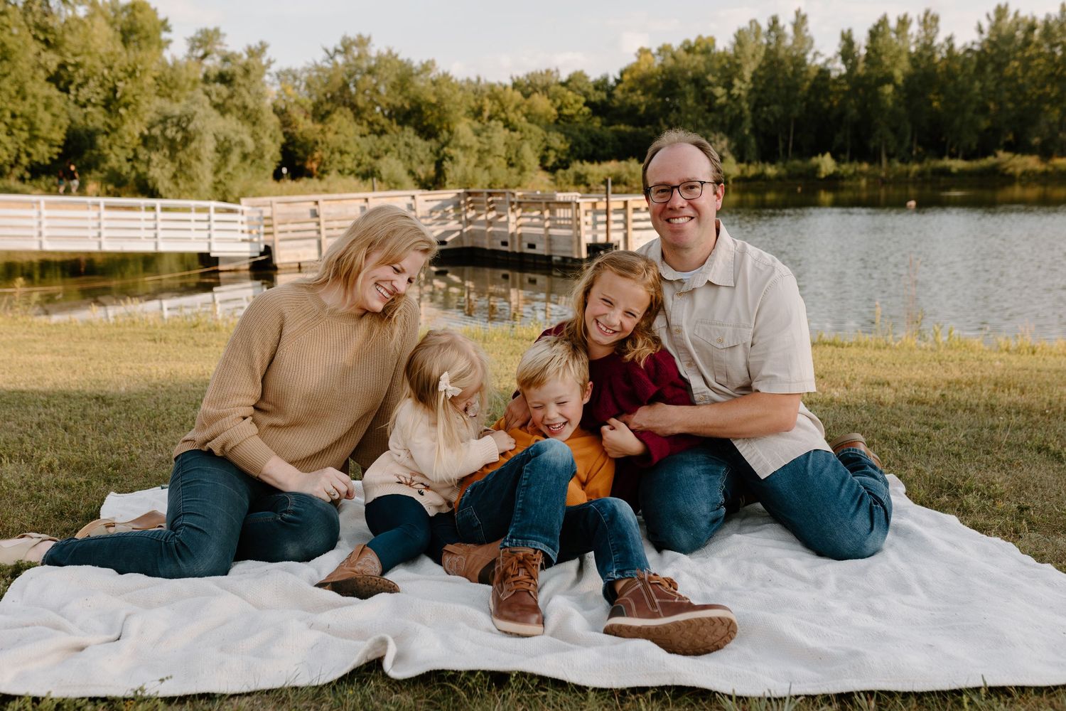 Family relaxing together on white blanket during outdoor photoshoot in sunny park setting.