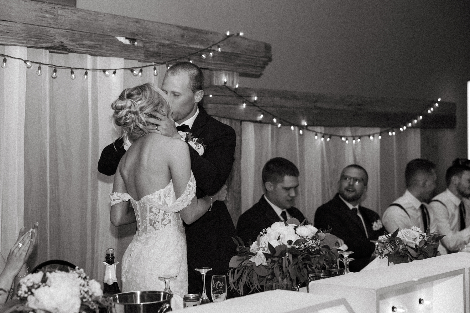 Bride and groom share a kiss at reception head table surrounded by wedding party in black and white.