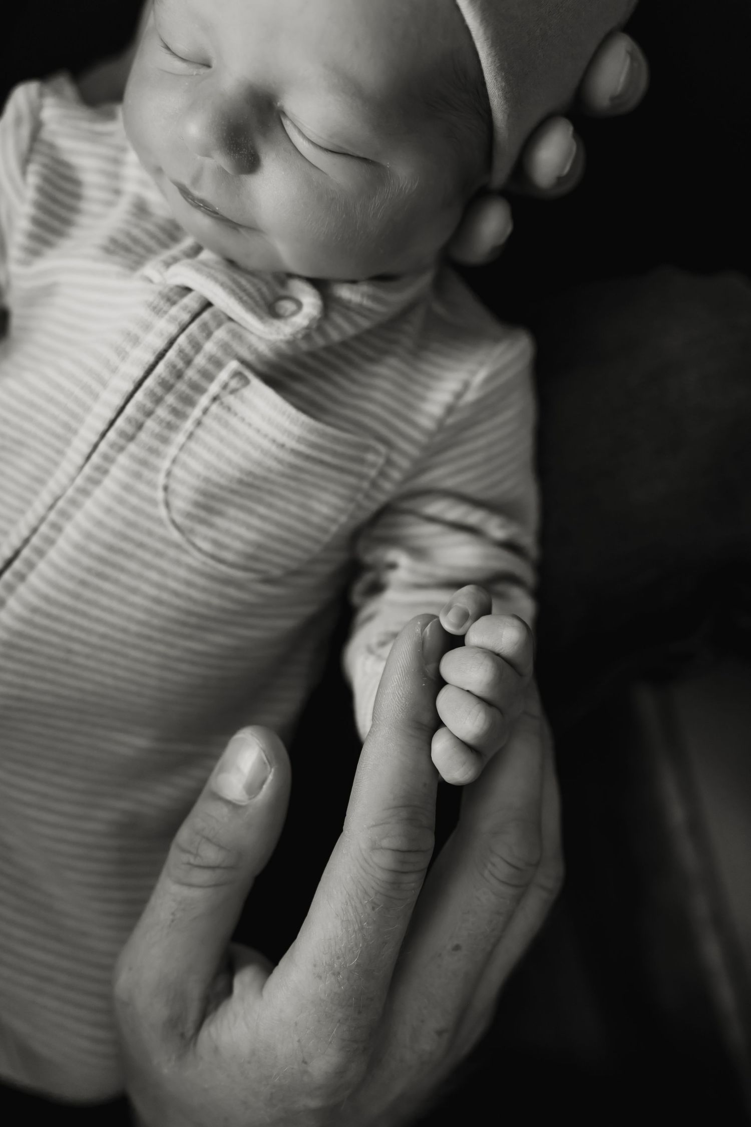 A sleeping newborn in striped clothing grasps an adult finger in a tender black and white portrait.