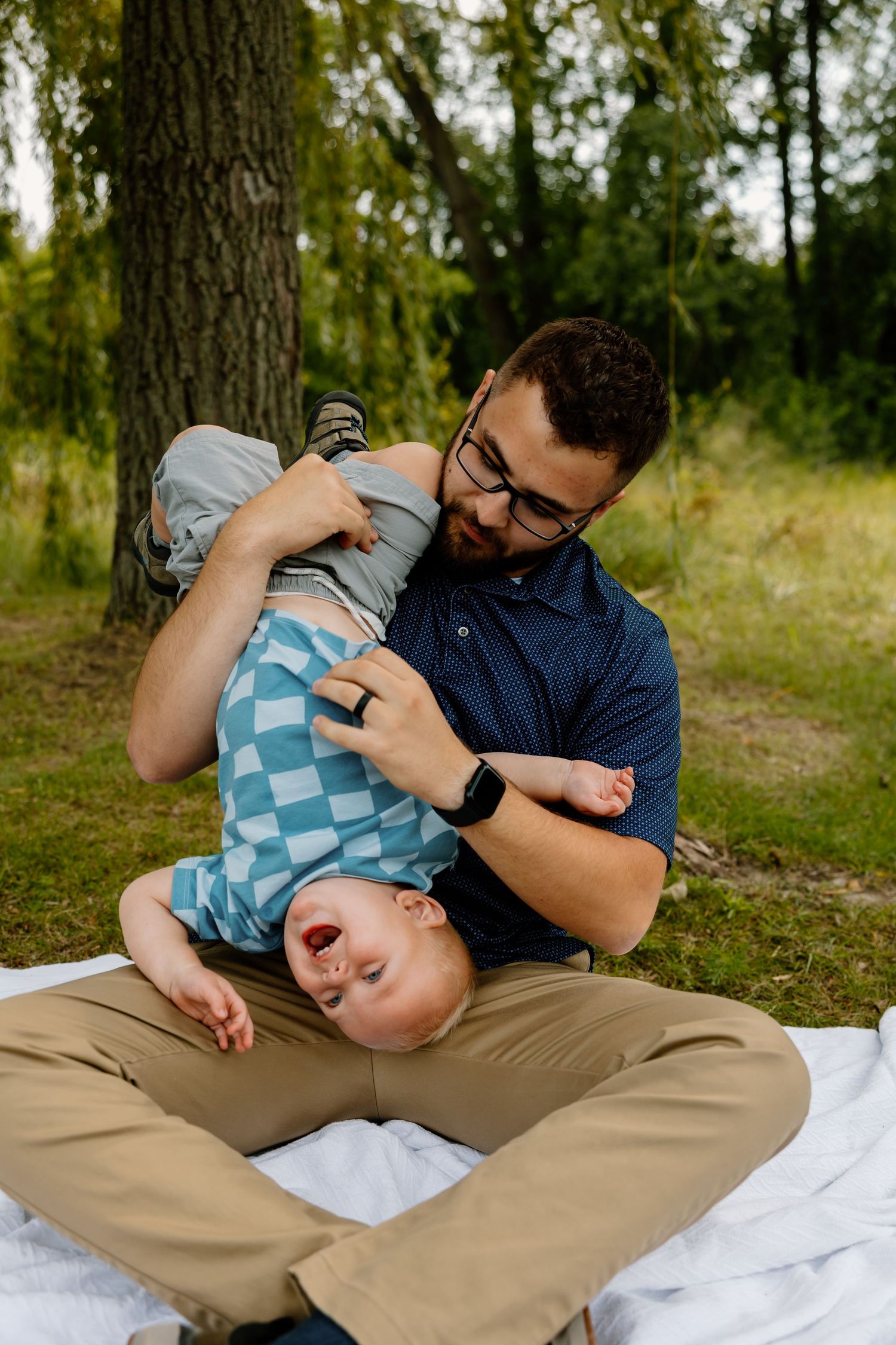 A playful moment of togetherness outdoors as a parent holds a laughing child upside down on a blanket in nature.