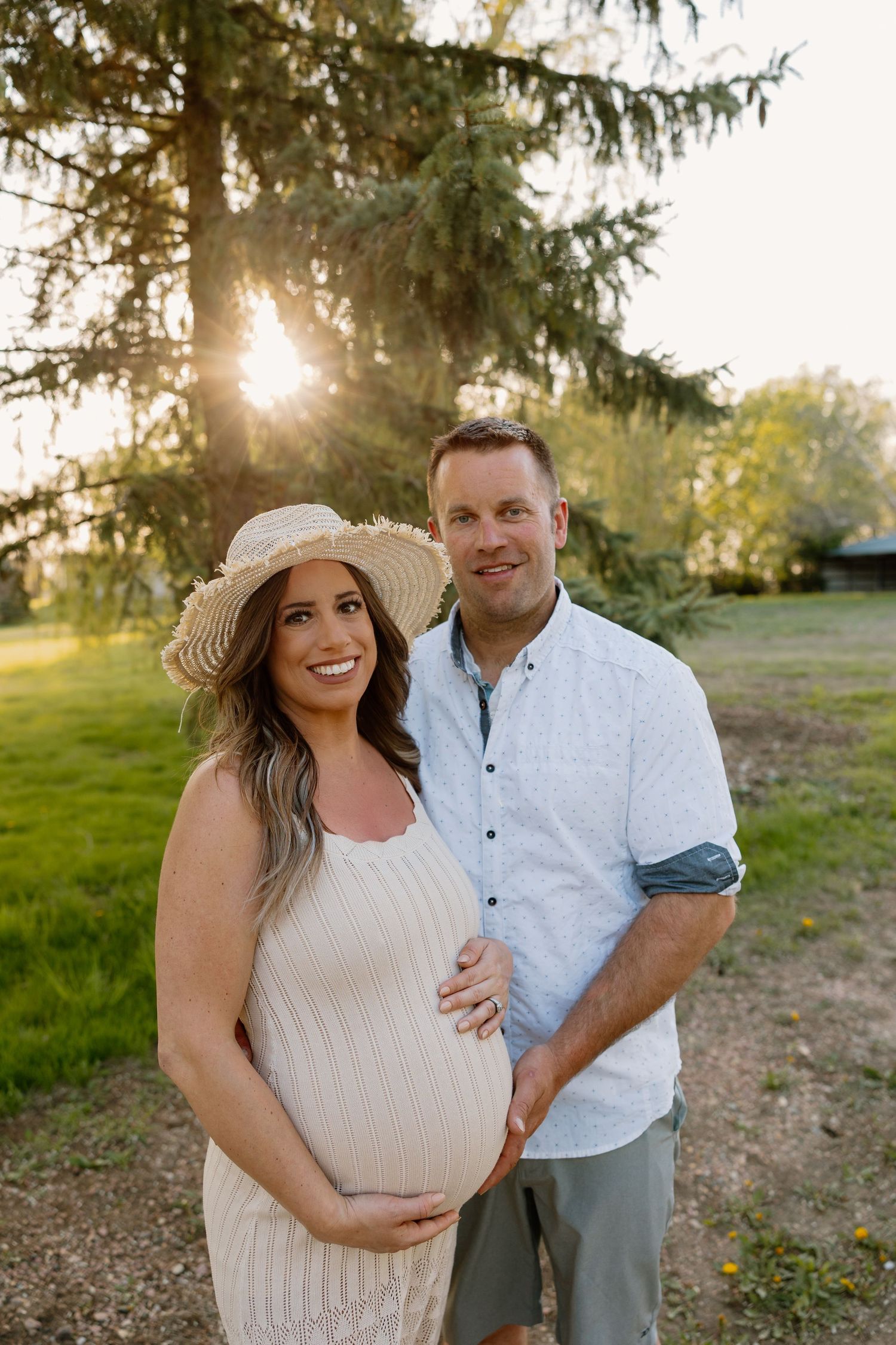 A couple stands together outdoors at sunset, with a pregnant person wearing a straw hat and white dress against evergreen trees.