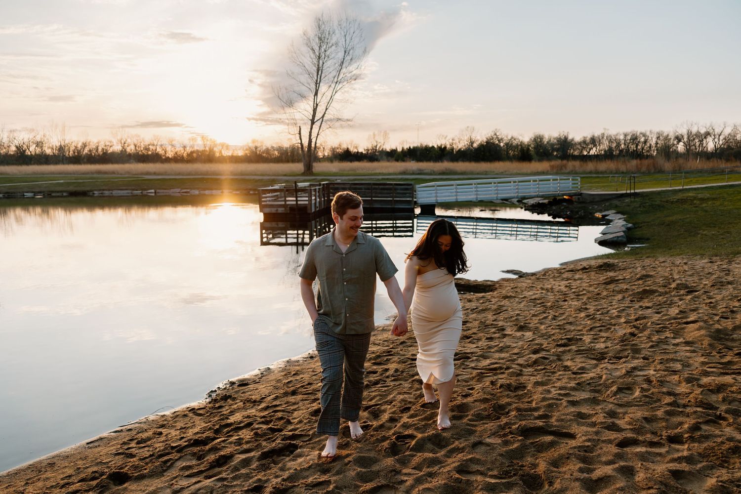 A couple walks along a sandy beach at sunset with a serene lake and bare tree in the background.