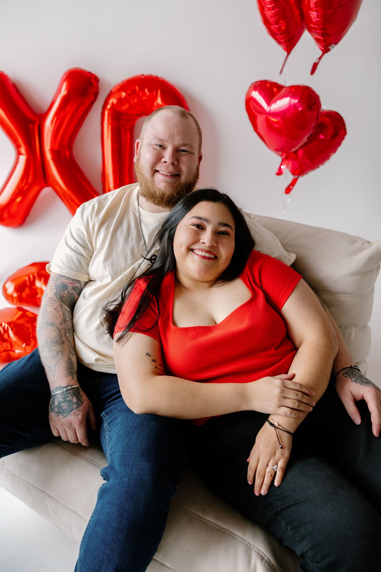 A couple relaxes on a white couch with festive red heart balloons and decorations for Valentine's Day.