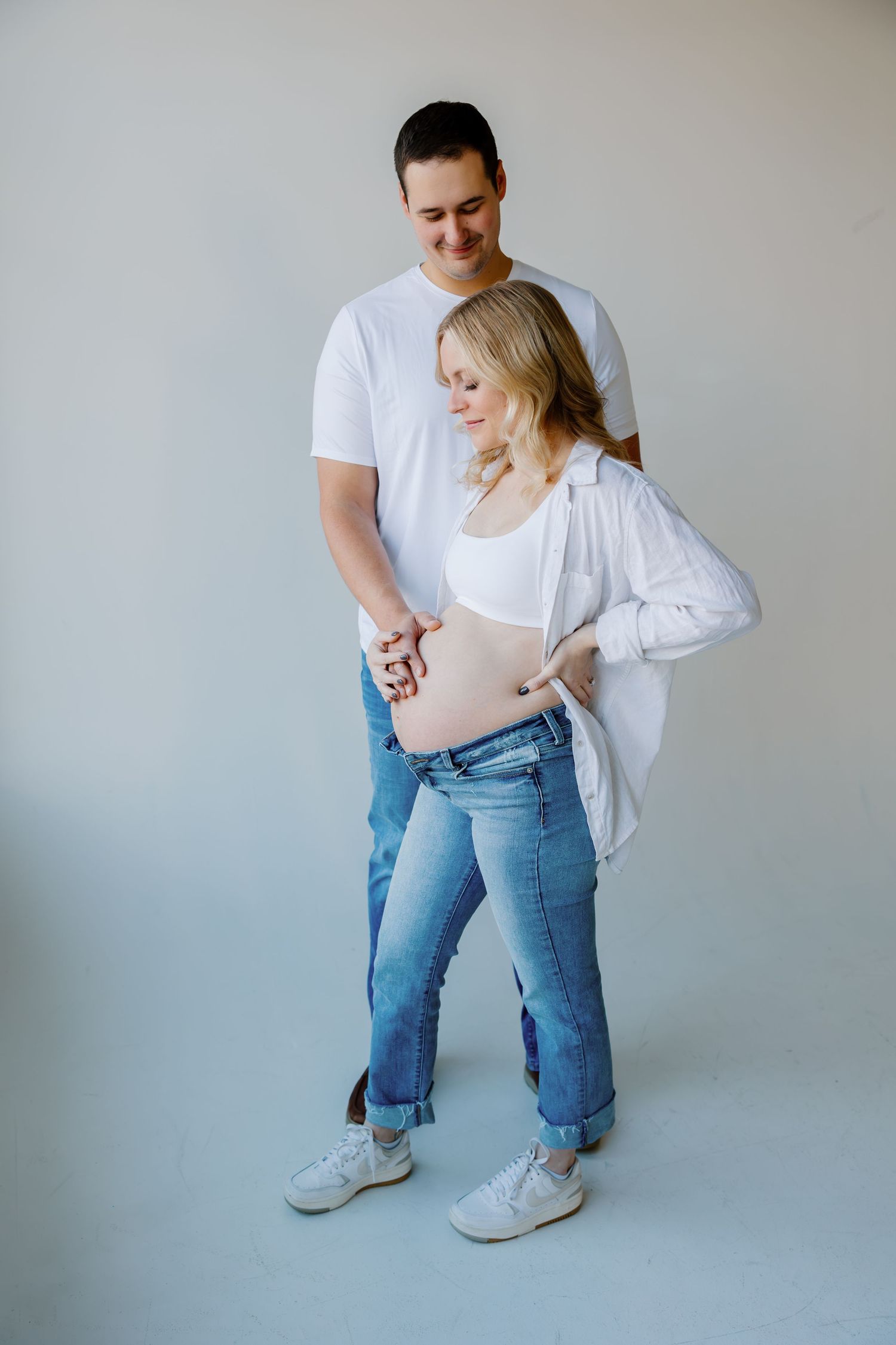 A couple in white shirts and jeans embraces during an intimate maternity photoshoot against a neutral backdrop. Riley Ro Photo Sioux Falls,