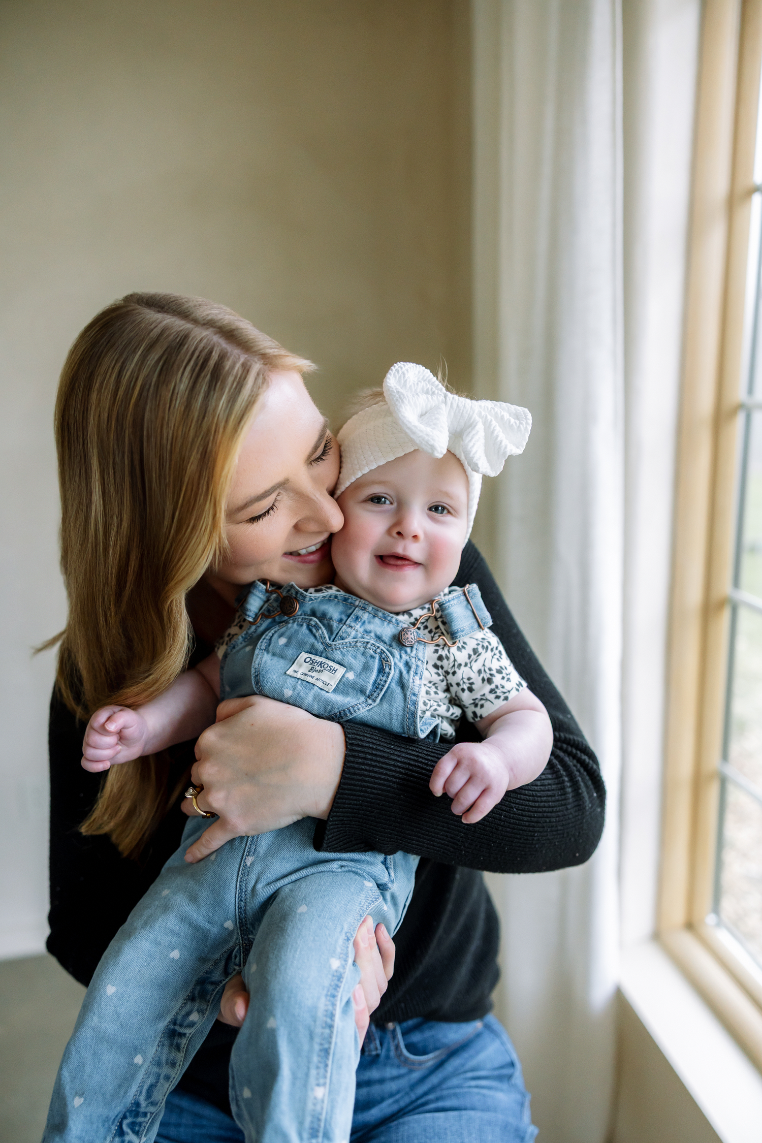 A mother tenderly kisses her smiling baby girl wearing a bow headband and denim overalls near a bright window.