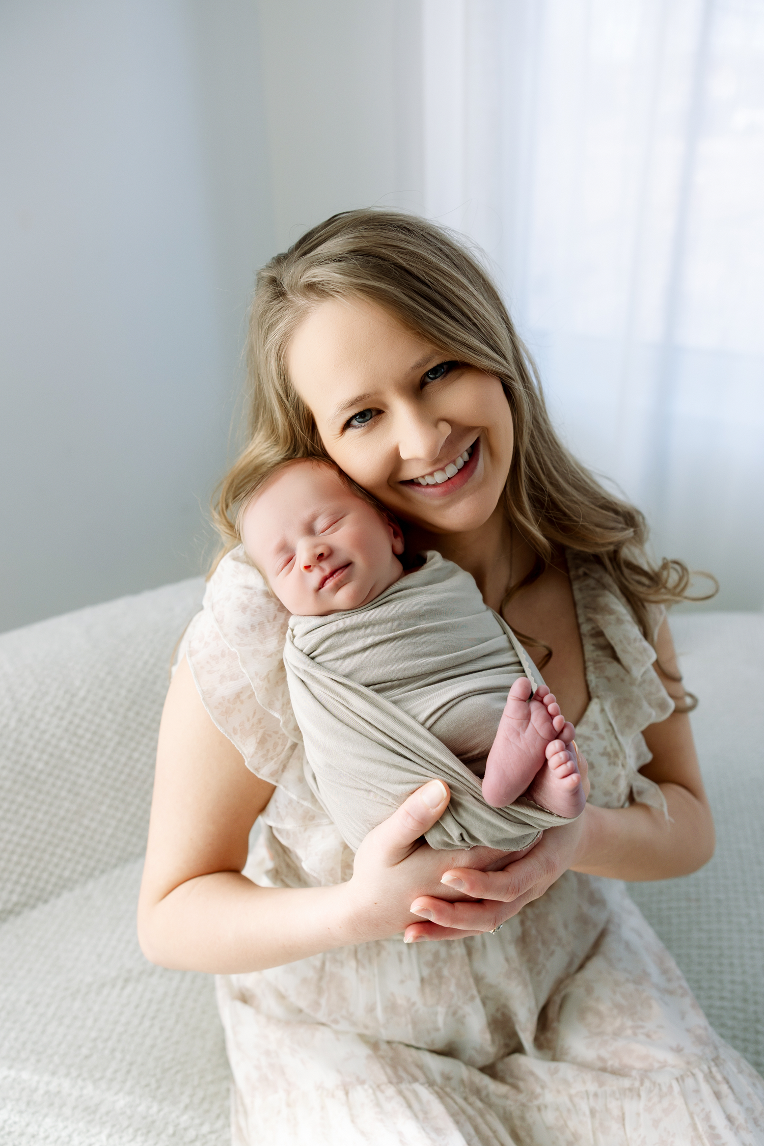 A parent and baby share tender moments together on a white couch, wrapped in a soft gray swaddle blanket.